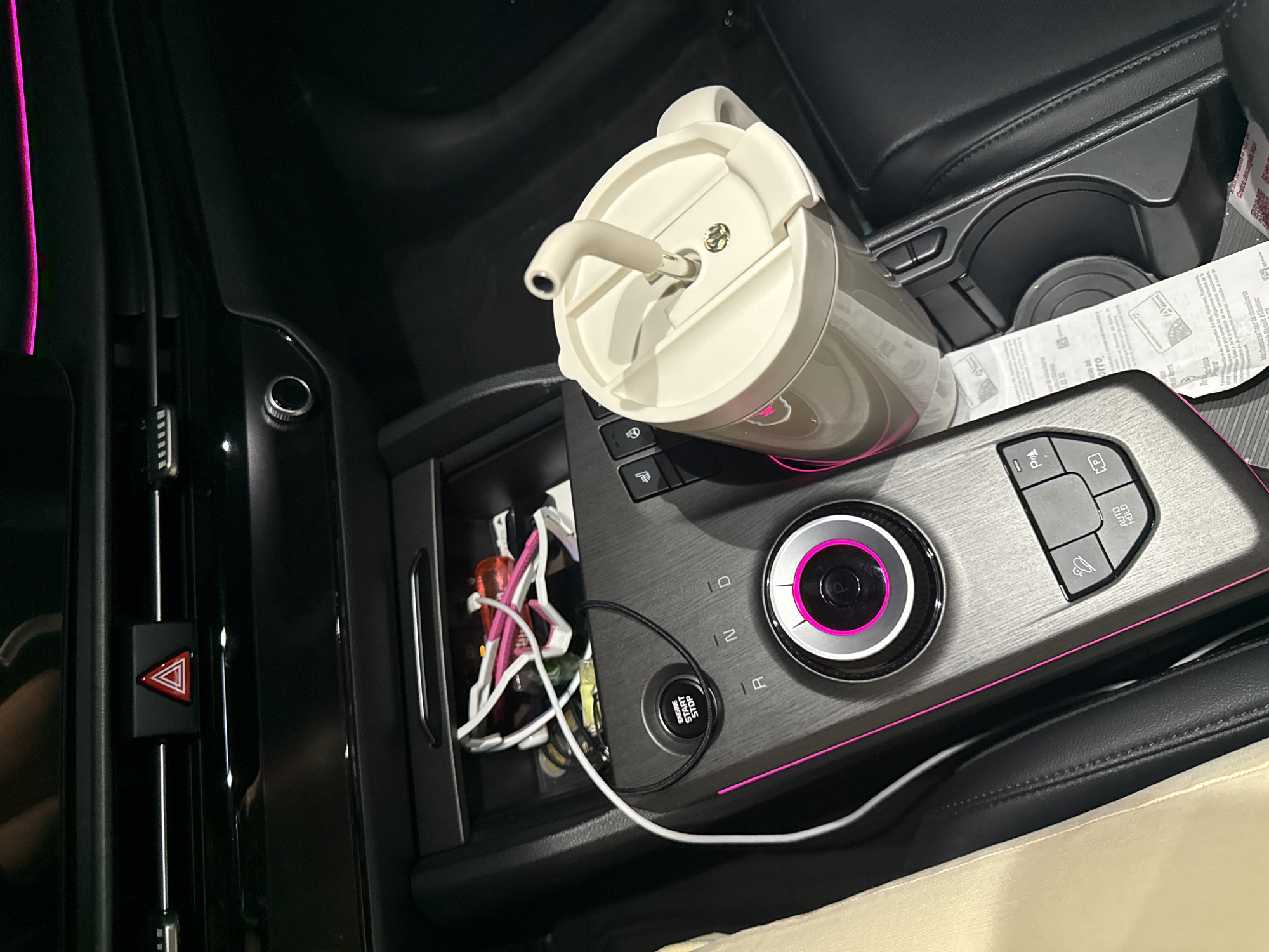 The image shows the interior of a car, specifically the center console area. A large, insulated cup with a straw is precariously placed near the cup holders. A gray control panel featuring buttons, a circular dial with pink accents, and an "Engine Start Stop" button is visible. An open storage compartment contains various items, including what appears to be cables and small objects. The car's hazard light button is on the left. Papers are in the cup holder.
The location of the image in San Bernardino Tlaxcalancingo, Mexico, cannot be determined from the visual data.