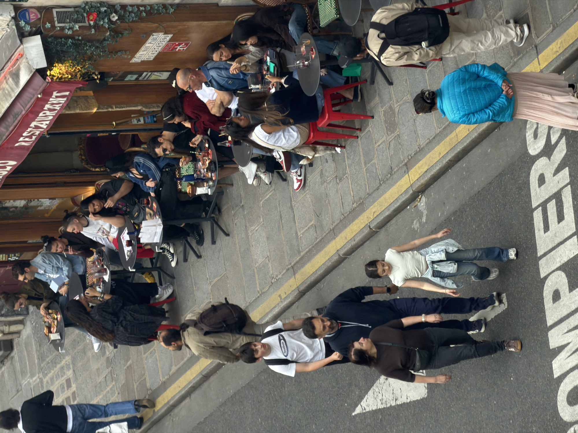 The image captures an outdoor cafe scene in Paris, France. Patrons sit at tables under the red awning of a restaurant, engaging in typical cafe activities. Outside the cafe's seating area, on the street marked with "COMPLÉTER" signage and directional arrows, a group of people lie on the ground, seemingly posing for a photo. The scene is captured from an elevated perspective, giving a comprehensive view of the interaction between the cafe and the street activity.