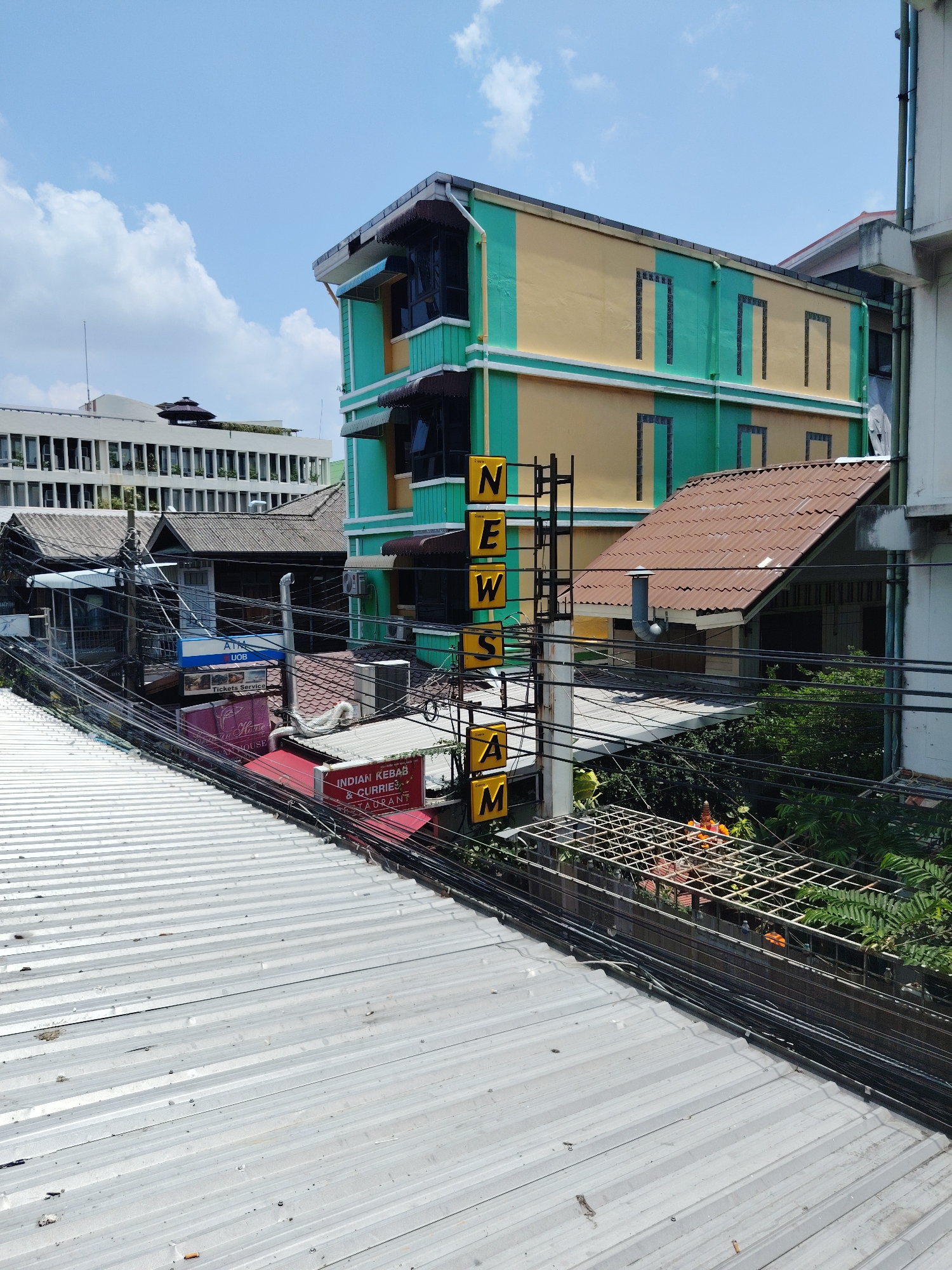 This photograph, taken in Bangkok, Thailand, presents an elevated view of a dense urban scene. In the foreground is a corrugated metal rooftop, leading the eye to a midground teeming with a mix of commercial and residential structures. A prominent vertical sign reading "NEWS AM," in yellow block letters, pierces the scene, indicating a media presence.
Other signage indicates the presence of an "Indian Kebab & Currie Restaurant" and an "ATM UOB," further anchoring the location as a commercial district. Overhanging wires add to the visual complexity. A building with a yellow and green façade, adorned with dark window awnings, stands out.
The background reveals additional buildings, including a modern white building with many windows. A variety of roof types, including tiled and corrugated, reflect the architectural diversity. Patches of vegetation are visible. The sky is partly cloudy. There are no people visible in this image.