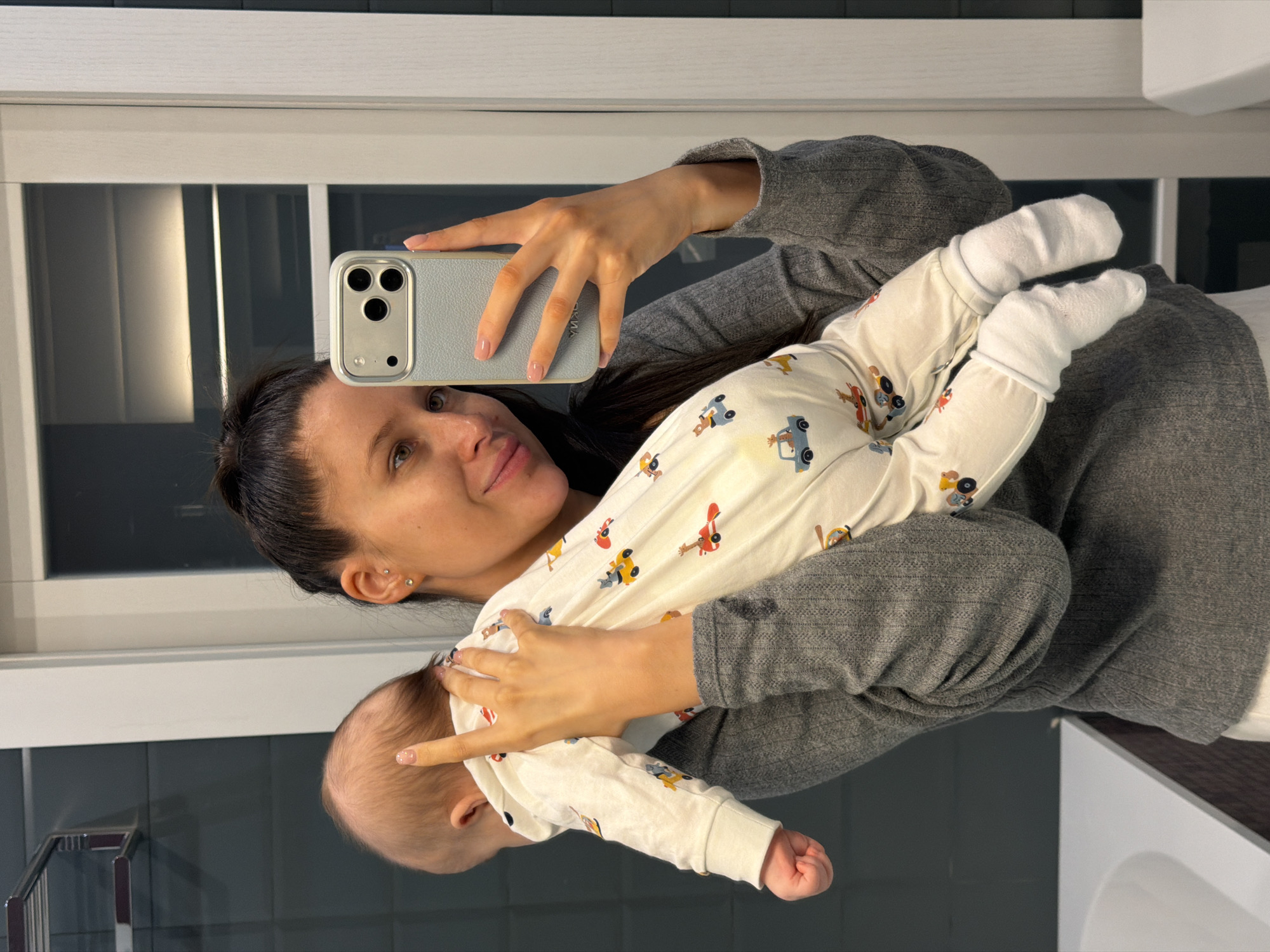 A woman is holding a baby and taking a selfie with a cell phone. She is wearing a gray long-sleeved shirt and has dark hair pulled back. The baby is wearing a white onesie with cartoon vehicle patterns and white socks. The background shows a bathroom setting with a window and gray wall. The location is Khimki, Russia.