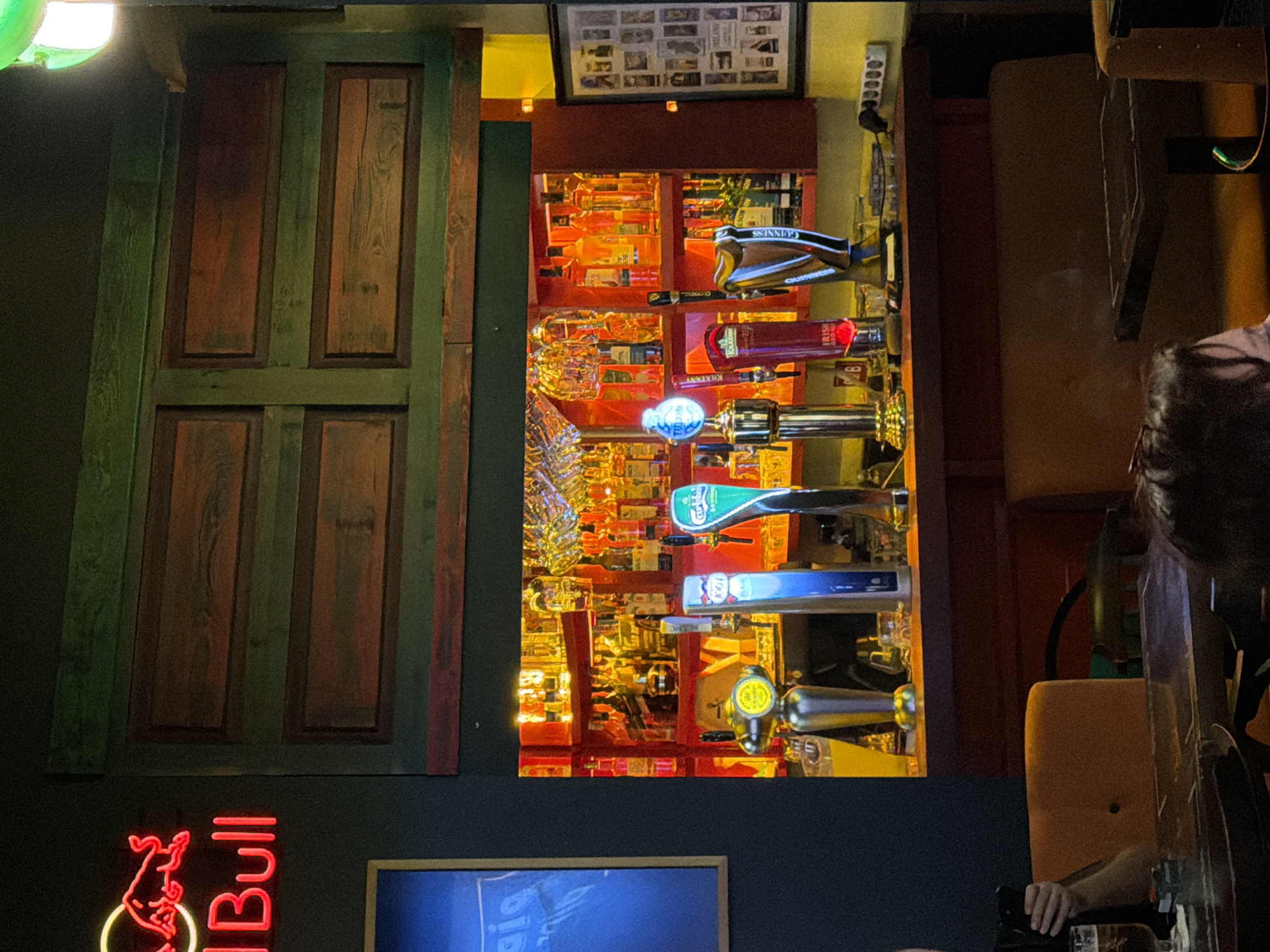 This image captures an interior scene of a bar. In the background, a well-stocked bar shelf is visible, illuminated by warm lighting, displaying numerous bottles of alcohol and hanging glassware. In the foreground, a row of beer taps stands prominently, including a Guinness tap. To the left, a neon red "Red Bull" sign is mounted on a dark wall. The lighting is dim, characteristic of a bar environment. The overall atmosphere suggests a commercial establishment focused on serving beverages. There is no information in the image that indicates the location is in Kadıköy, Turkey.