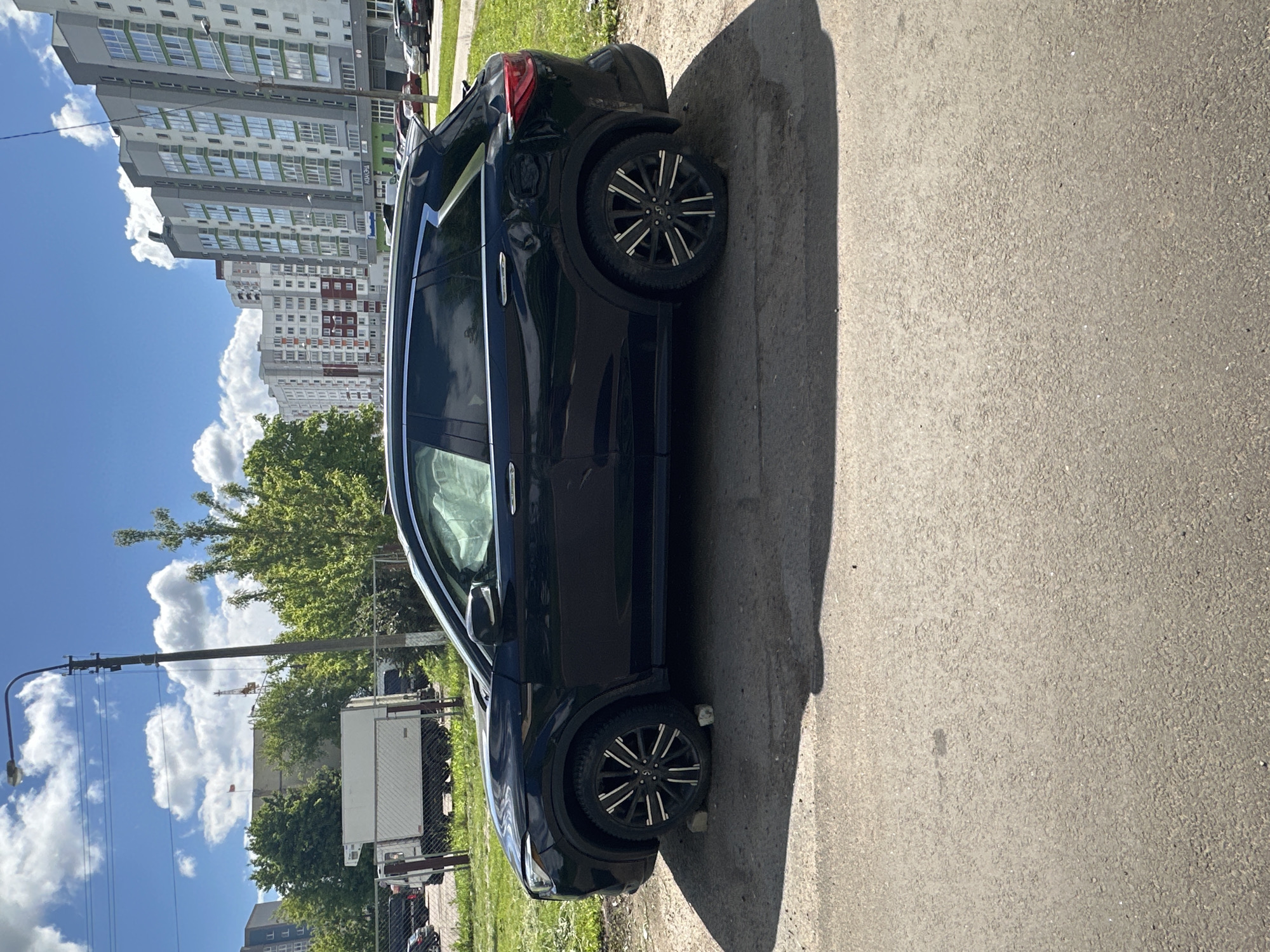 The image shows a black SUV parked on the side of a road. Buildings are visible in the background, suggesting an urban environment. Other parked cars and a white truck are also present in the background, indicating a street with parking. A lamp post is visible in the left part of the image. Minsk, Belarus, is a possible location given the architectural style of the buildings.