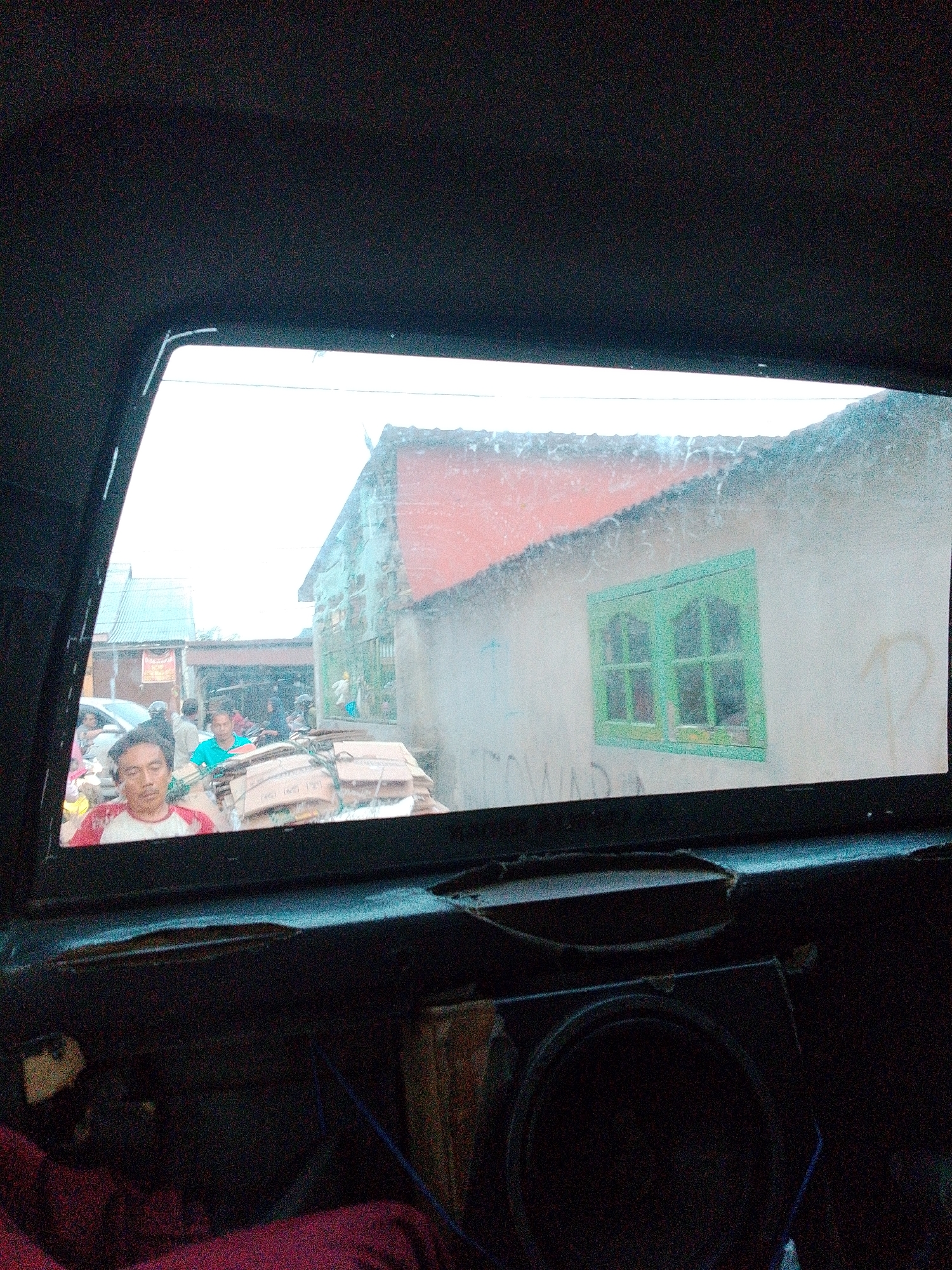 The image depicts a view through the rear window of a vehicle, looking out onto a street in Bandar Khalipah, Indonesia. A man, wearing a red and white shirt, is visible in the foreground on the left side of the window, his face turned slightly towards the camera. Behind him, more people are visible on motorbikes and on foot, some carrying bundles of cardboard boxes. The street appears to be lined with buildings, one of which has a prominent green-framed window. The scene suggests a busy, possibly market-like environment. The interior of the vehicle is visible at the bottom of the frame, showing a speaker. The overall lighting suggests an overcast day.