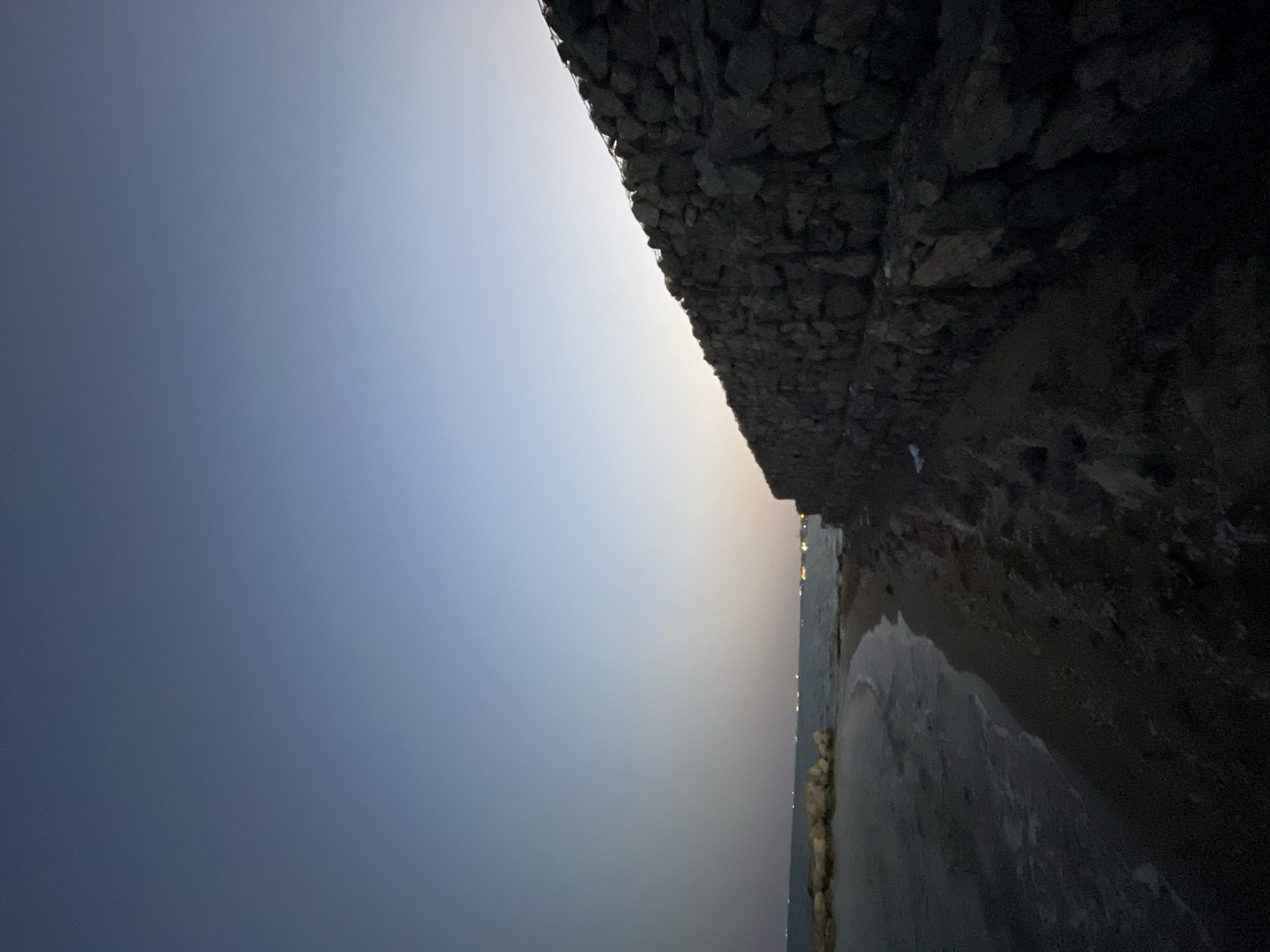 An outdoor, wide-angle shot captures a beach scene at dusk, looking out towards the sea. A prominent gabion wall, constructed of stacked stones, runs diagonally across the right side of the frame, creating a stark contrast with the natural elements. The water is calm, with gentle waves lapping at the sandy shore. In the distance, faint lights suggest a settlement or possibly boats. The sky transitions from a deep blue at the top to a lighter hue near the horizon. The overall impression is one of a quiet, serene evening by the coast. This scene is located in Trikomo, Northern Cyprus.
