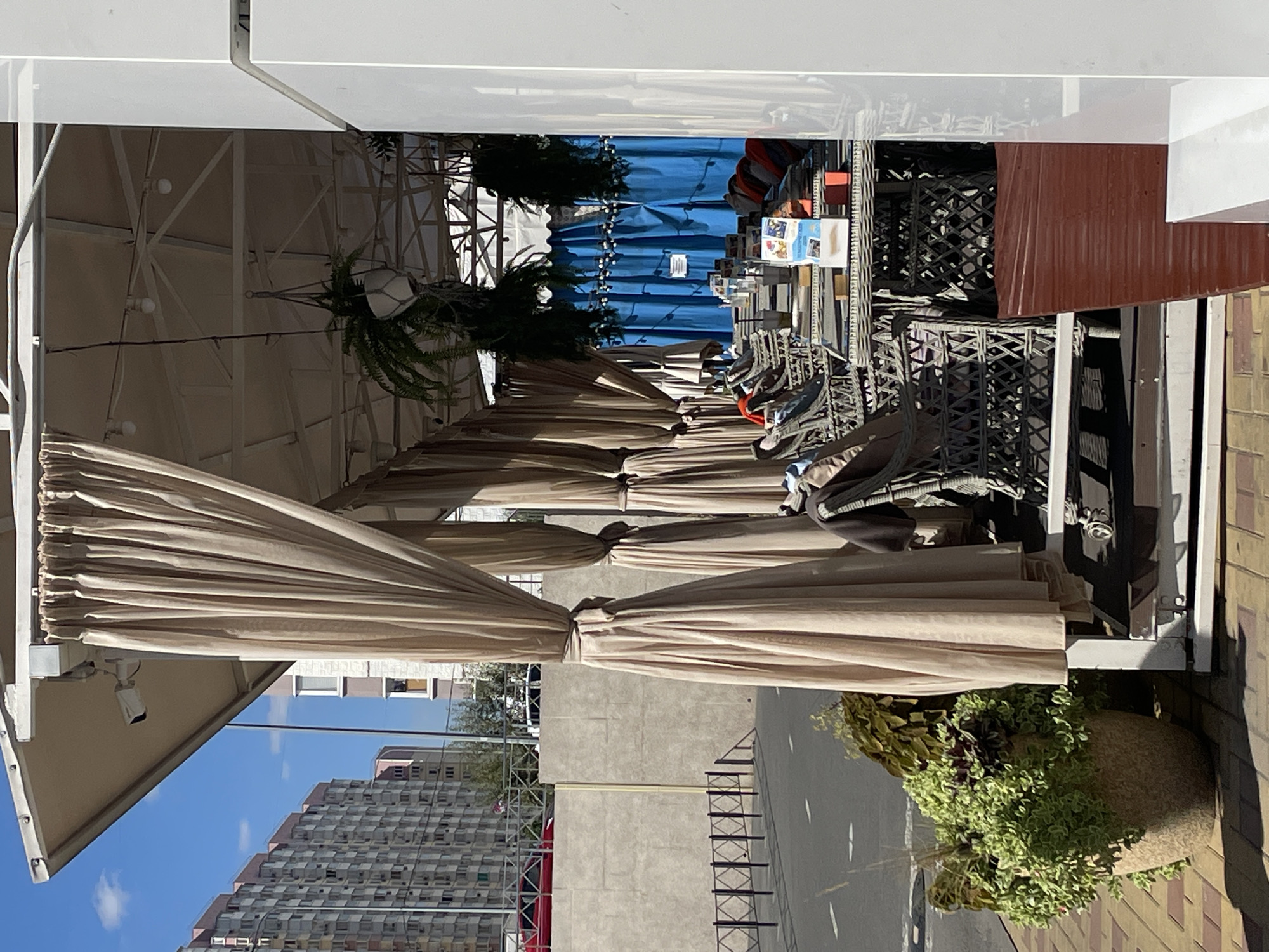This image depicts an outdoor cafe seating area in Rostov-on-Don, Russia, on a sunny day. In the foreground, a large potted plant with lush green foliage sits on a tiled walkway. Behind the plant, a white outdoor structure with beige curtains creates a shaded area. Tables and wicker chairs are arranged within this area, some with blankets or cushions. The tables are set with various items, including glasses, coasters, and a brochure. In the background, a concrete wall separates the cafe from a road with white lane markings. Beyond the wall, tall apartment buildings are visible under a clear blue sky with scattered clouds. The overall scene suggests a peaceful, outdoor dining or relaxation space.