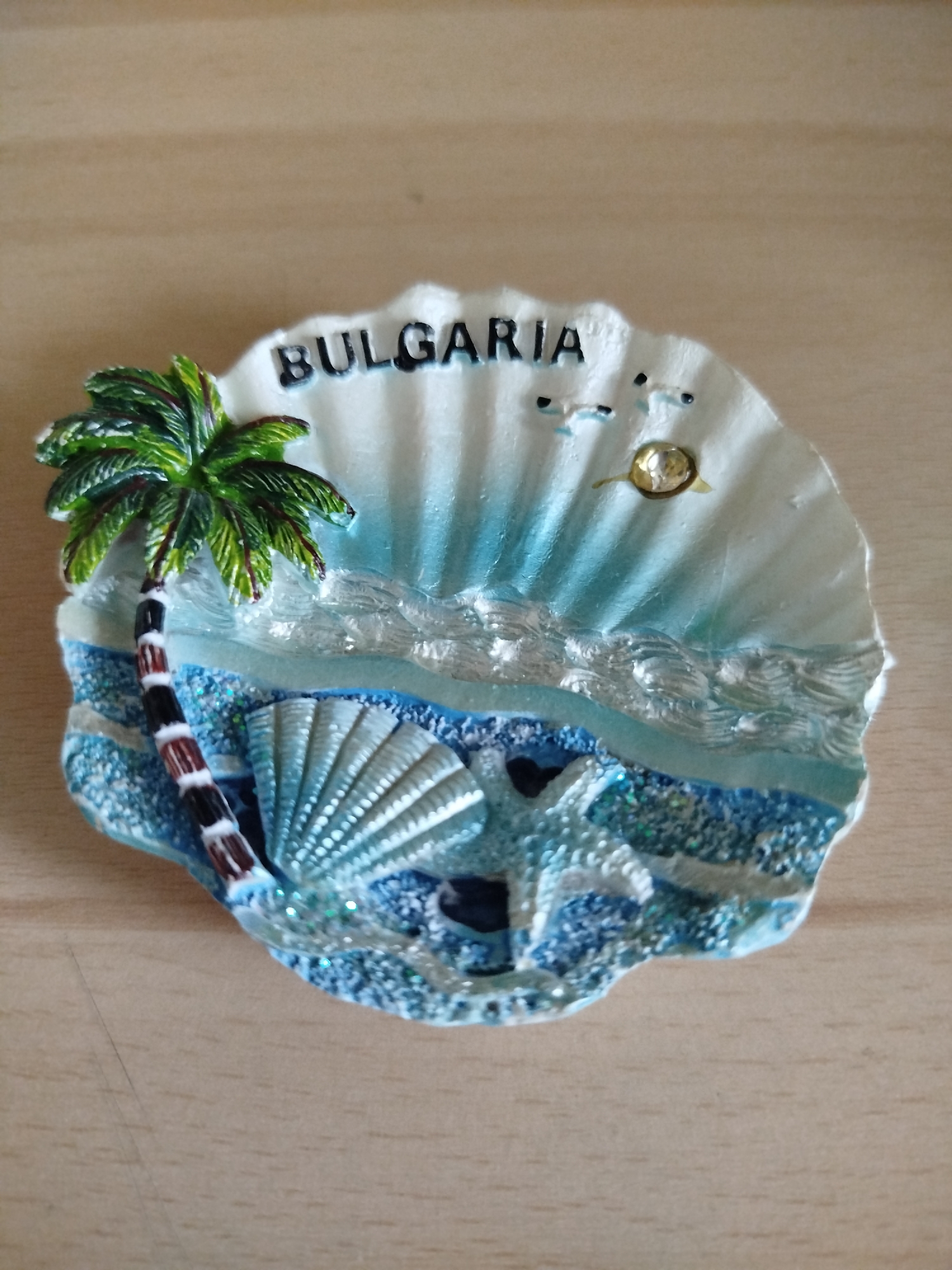 The image shows a seashell-shaped souvenir magnet from Bulgaria. The magnet features a palm tree on the left, a seashell and starfish in the center, and the word "BULGARIA" at the top. It includes decorative elements representing the sea, such as waves and blue glitter. Given the coastal theme, the souvenir likely originates from a seaside city, such as Varna, Bulgaria.