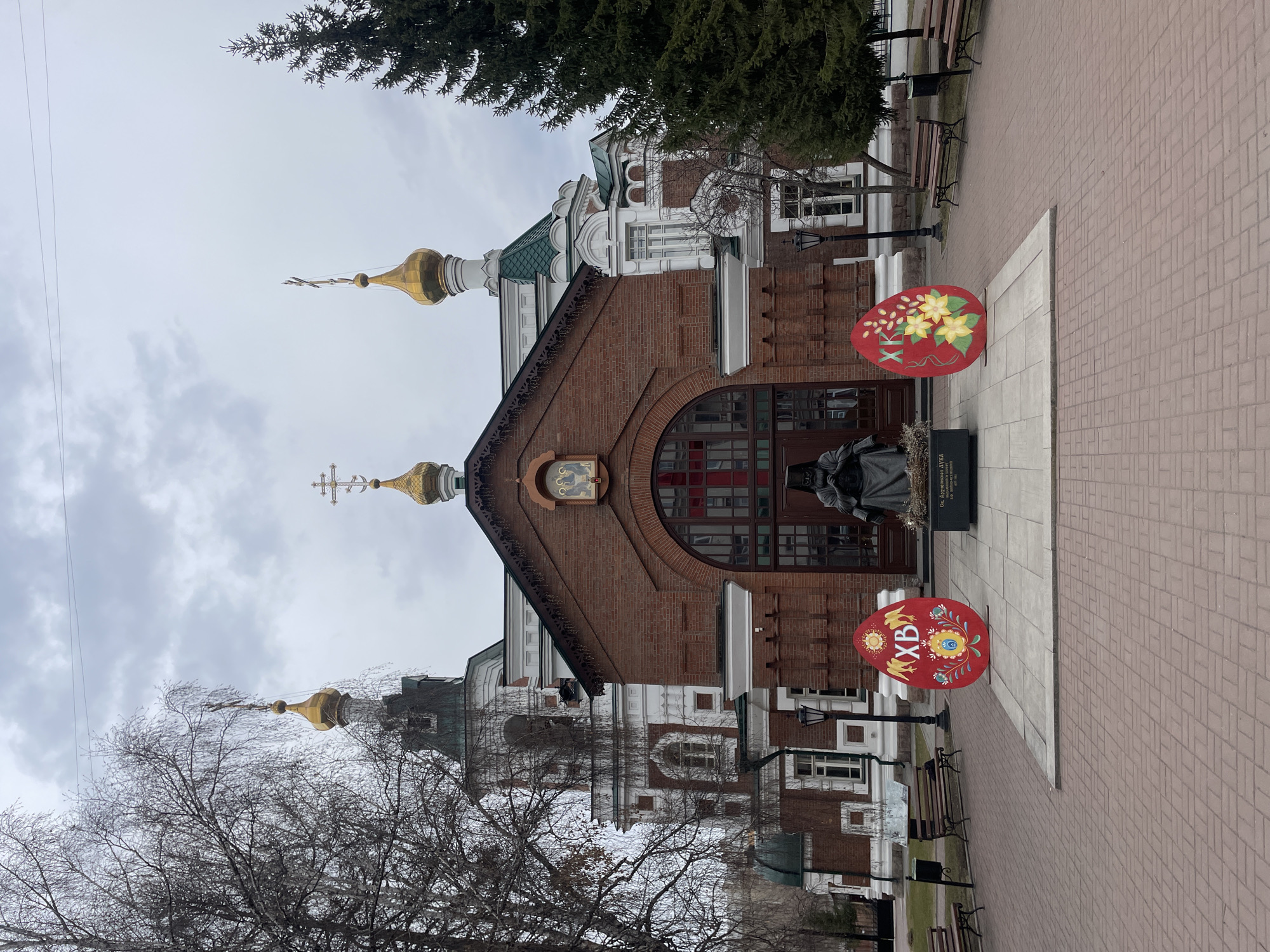 An Orthodox church with distinct Russian architectural features stands prominently in the center of the image, under an overcast sky. The main body of the church is constructed from red brick, featuring white decorative elements, arched windows, and a green roof. Two golden onion domes, each topped with a cross, rise from the structure; a smaller one over the entrance and a larger one further back.
In front of the church entrance, a dark-colored statue of a robed figure stands on a stone pedestal. While the full text on the pedestal is not clearly legible from this distance, the partial text visible on the crop reads "СВ. ПРЕПОДОБНЫЙ СЕРАФИМ САРОВСКИЙ" (Saint Reverend Seraphim of Sarov).
Flanking the statue are two large, flat, egg-shaped decorations, painted in red with elaborate colorful patterns. The lower egg clearly displays the Cyrillic letters "ХВ", signifying "Christ is Risen," a common Easter greeting. The upper egg features floral designs in yellow and green. These decorations suggest an Easter season setting.
The area around the church is a paved square with light-colored bricks. Several dark benches and street lamps are visible along the paved paths. Bare trees with intricate branches line the foreground and background, indicating a cold season like late winter or early spring, while an evergreen fir or spruce tree is also visible on the right. The sky is uniformly gray and cloudy, suggesting daytime but likely a cool or chilly environment in Krasnoyarsk, Russia. No specific activity or people are present, lending a quiet atmosphere to the scene.