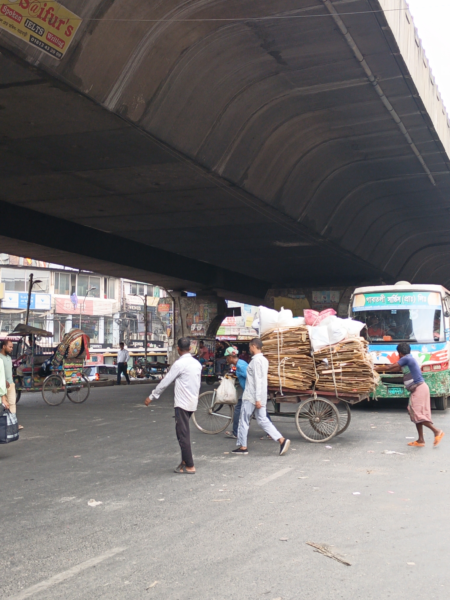 The image shows a street scene in Dhaka, Bangladesh, under a large overpass. The overpass is made of concrete. A sign for "Saifur's" is affixed to the structure.
A three-wheeled cart loaded with bundled cardboard and white sacks is being pulled through the scene. A bus with the name "গাবতলী সার্ভিস (প্রাঃ) লিঃ" (Gabtoli Service (Pvt.) Ltd.) is in the background. People are walking around the cart and the bus. There are also multiple bicycle rickshaws on the left.
The buildings are old and crowded with advertisements and signs in the background. The street appears to be busy with traffic.
