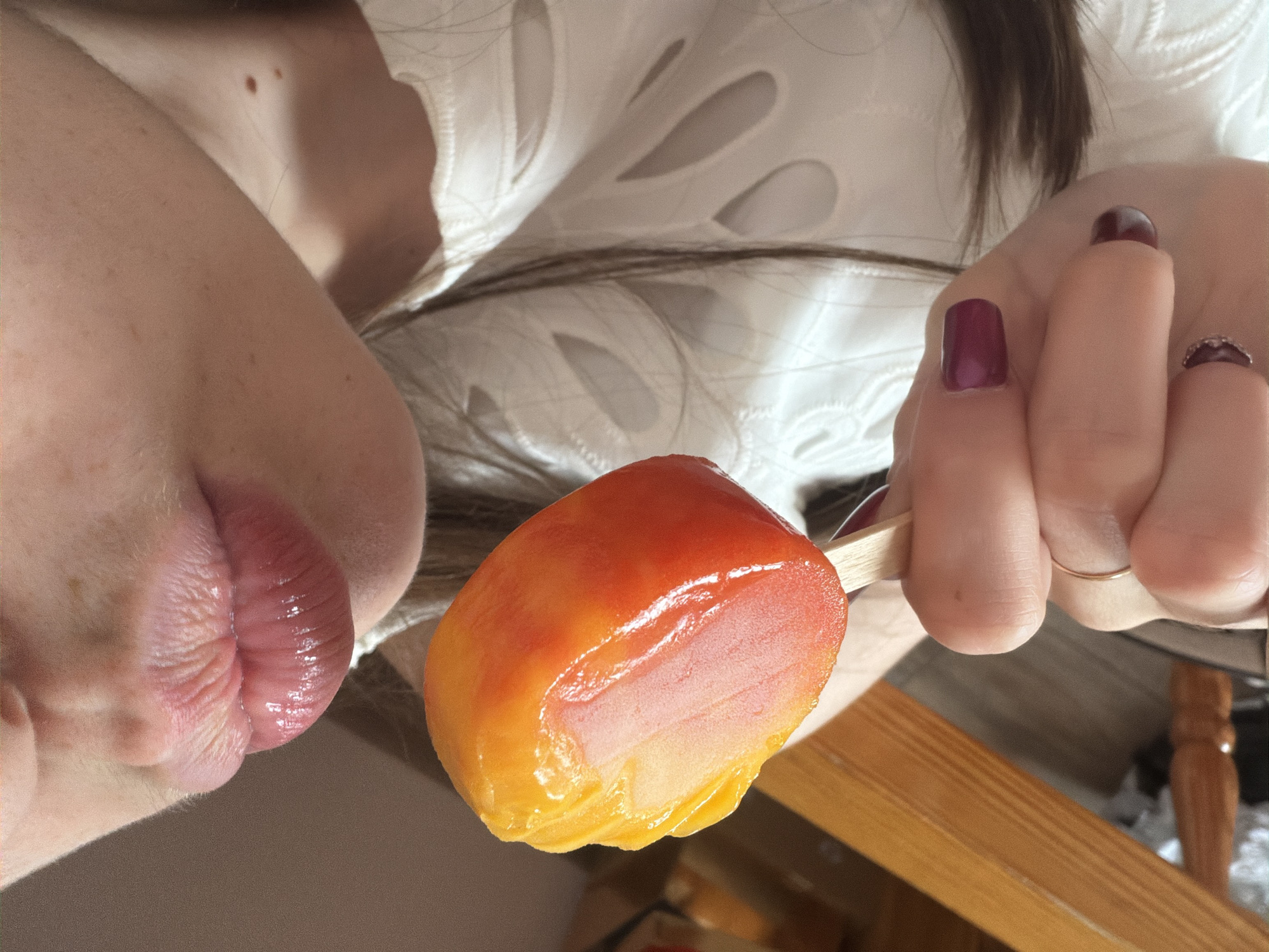 A close-up, eye-level indoor shot depicts the lower portion of an individual's face, specifically the chin and lips, and one hand holding a popsicle. The lips are full, glossy, and slightly pursed, indicating an action of eating or about to eat. The skin appears fair with several small moles or freckles visible on the cheek and chin area. Long, dark brown hair cascades over the individual's right shoulder, partially obscuring a white garment with distinct, intricate cut-out patterns.
The individual's right hand holds a light wooden stick embedded in a brightly colored popsicle. The popsicle is ovate, exhibiting a gradient from vibrant yellow and orange at the top to a deeper reddish-orange at the bottom, with a lighter pinkish-orange interior visible where a portion has been consumed or is exposed. The surface of the popsicle appears wet and glistening. The fingernails are painted a deep plum or burgundy color, and a thin, plain gold ring is worn on one finger.
In the background, to the left, a section of light brown wooden furniture, possibly a chair arm or table leg, is visible. The remainder of the background is out of focus, showing muted, warm tones characteristic of an indoor setting. The specific location within the city of Varna, Bulgaria, cannot be determined from the visual information provided.