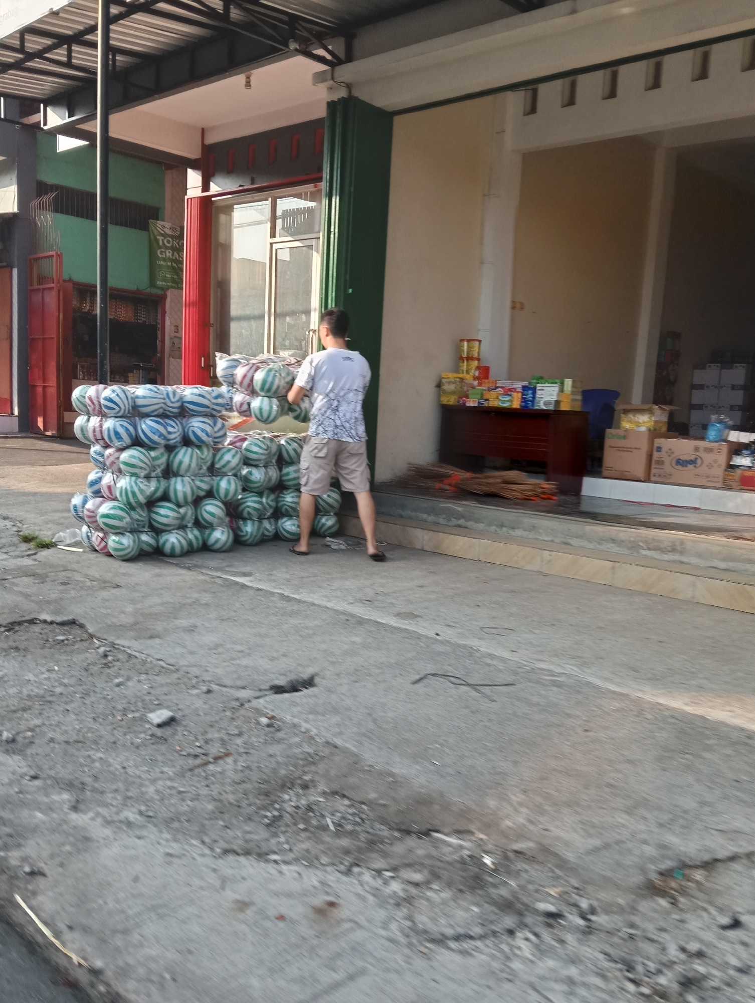 The image depicts a street scene in Grabag, Indonesia. A man wearing a white t-shirt and khaki shorts stands in front of a large stack of what appear to be inflated balls, possibly for sale or delivery. The balls are wrapped in plastic and are a mix of green, white, and red colors. To the right of the stack is a storefront with a counter displaying various products, including what look like canned goods and boxes. A pile of brooms rests on the ground in front of the counter. In the background, there are more storefronts and structures, suggesting a commercial area. The ground is paved with concrete and shows some signs of wear and tear. The lighting indicates it is daytime, possibly late afternoon, given the shadows.