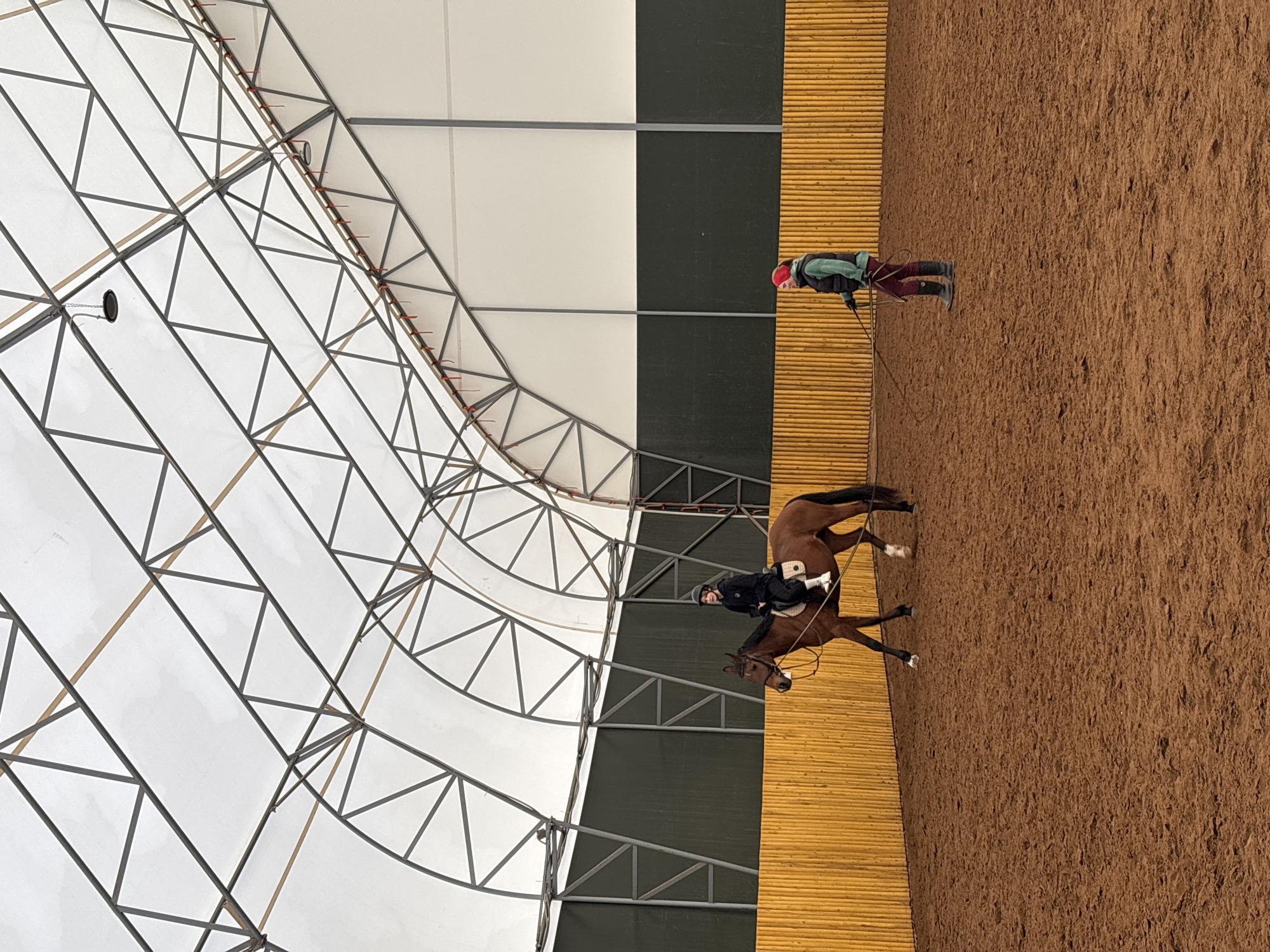 The image depicts an indoor equestrian arena. A person is riding a brown horse within the arena. Another person stands along the edge of the riding area, holding a long rope. The arena has a tan-colored dirt floor and a tall wooden wall on one side. The arena's ceiling is composed of a white canopy supported by a steel frame. Based on information available, the location is in the city of Русский Вожой, Russia.