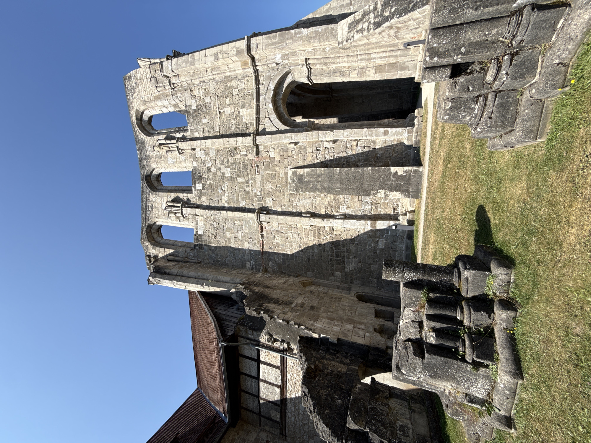 The image shows the exterior of the Walkenried Abbey in Walkenried, Germany. The structure is partially ruined, with large stone walls and arched windows visible. In the foreground, there are several large stone fragments and pillars on a grassy area. To the left, a section of the abbey is intact, featuring a red tiled roof and timber-framed walls. The sky is clear and blue.
