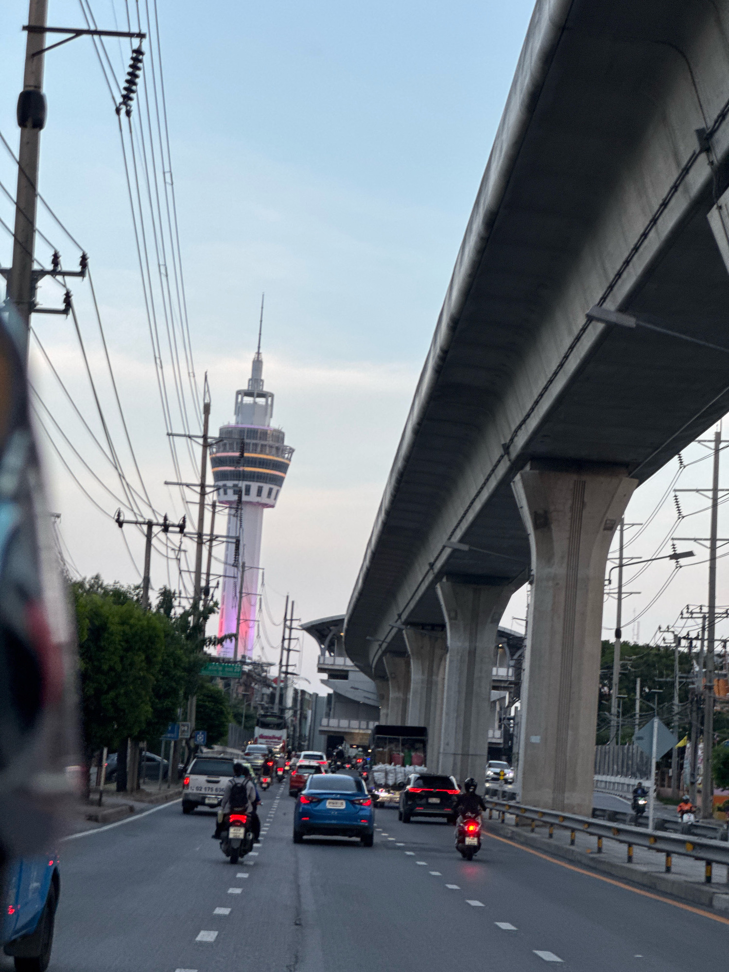 The image captures a street scene in Samut Prakan, Thailand, with a prominent tower in the background, illuminated with colorful lights. The sky is clear and appears to be late afternoon or early evening. A concrete elevated structure, likely part of a transportation system like an expressway or monorail, dominates the right side of the frame, supported by large pillars.
The road in the foreground is moderately busy with vehicular traffic, including cars and motorcycles. Several vehicles are visible, such as a blue sedan, a white SUV, and multiple motorcycles. People are riding on the motorcycles, some wearing helmets. The road has lane markings and is bordered by a guardrail on the right.
Utility poles with numerous electrical wires are scattered throughout the scene, a common feature in urban environments. Trees and some buildings can be seen along the side of the road, interspersed with the infrastructure. The overall impression is of a bustling urban environment on the outskirts of a city, with a notable landmark in the distance.