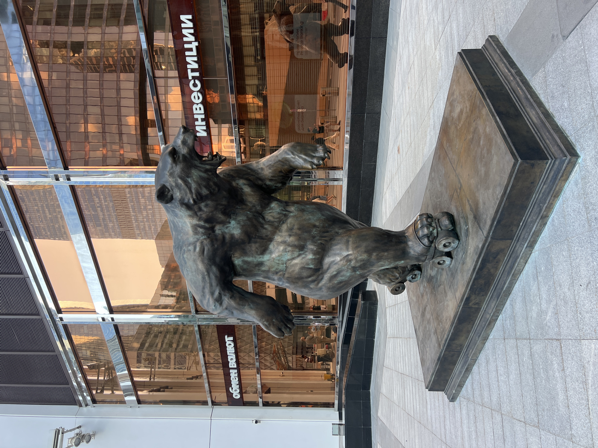 A bronze sculpture of a bear on roller skates stands on a rectangular base in front of a modern building with large, reflective glass windows. The building's facade is tinted amber, reflecting the sky and surrounding architecture. White text in Cyrillic, reading "ОБМЕН ВАЛЮТ" (Currency Exchange) and "ИНВЕСТИЦИИ" (Investments), is visible on the building. The ground is paved with light gray square tiles. In the reflection of the windows, other buildings and people can be seen. The lighting suggests it is daytime, possibly late afternoon due to the warm tones in the reflections. The sculpture is positioned centrally in the frame, facing slightly to the viewer's left, with its mouth open in a roar. The overall scene depicts an outdoor urban environment, likely a financial district given the building's signage. This sculpture is located in front of the Sberbank building in the city of Khimki, Russia.
