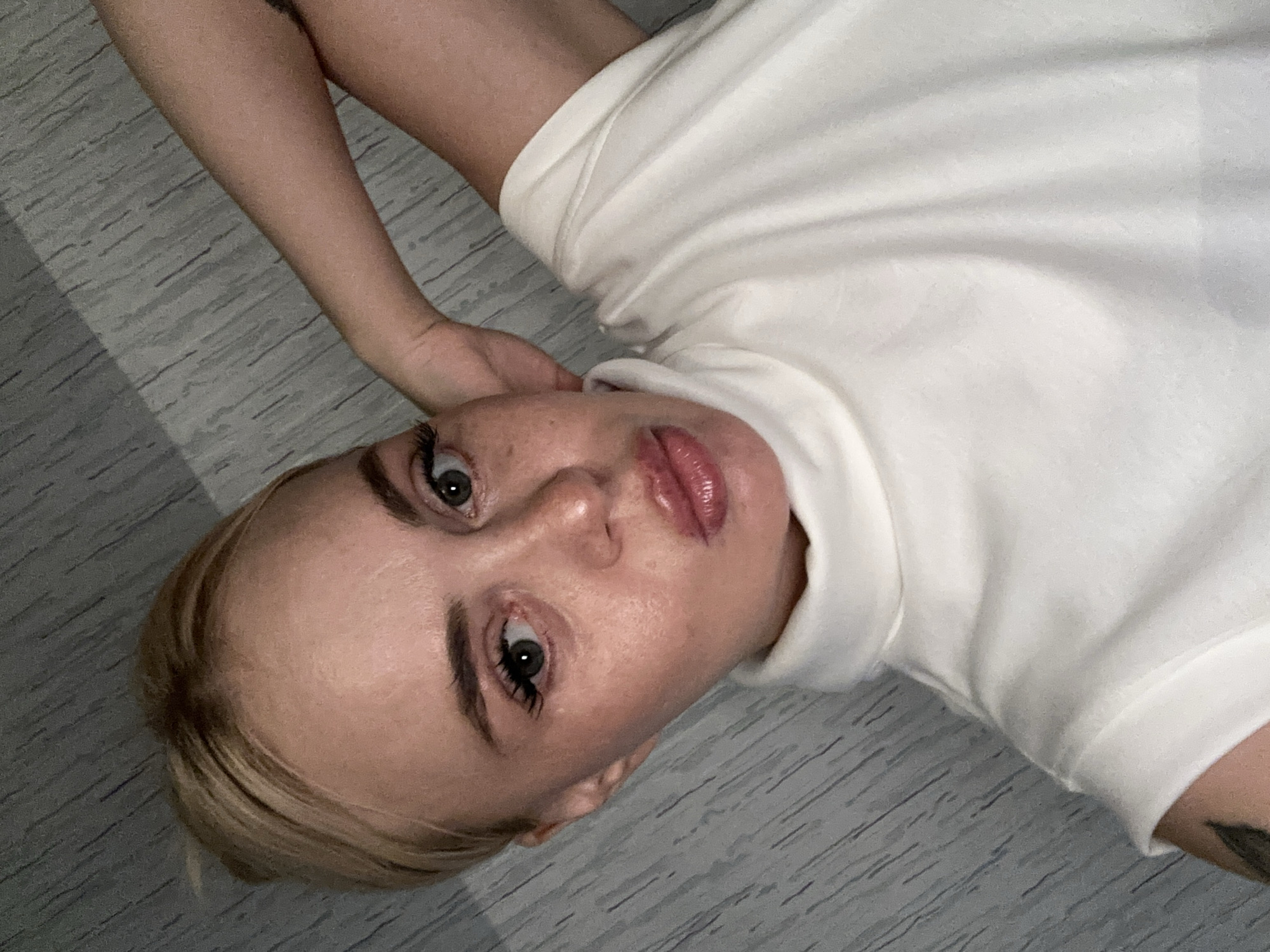 A close-up, high-angle, indoor selfie of a fair-skinned, blonde woman is depicted. She is positioned with her head tilted slightly, looking directly at the camera, with her left arm bent and resting behind her head. The woman has prominent eyebrows, fair skin with freckles, green eyes, and plump lips. She is wearing a white, high-necked shirt. The background consists of a gray, textured wall with diagonal lines.