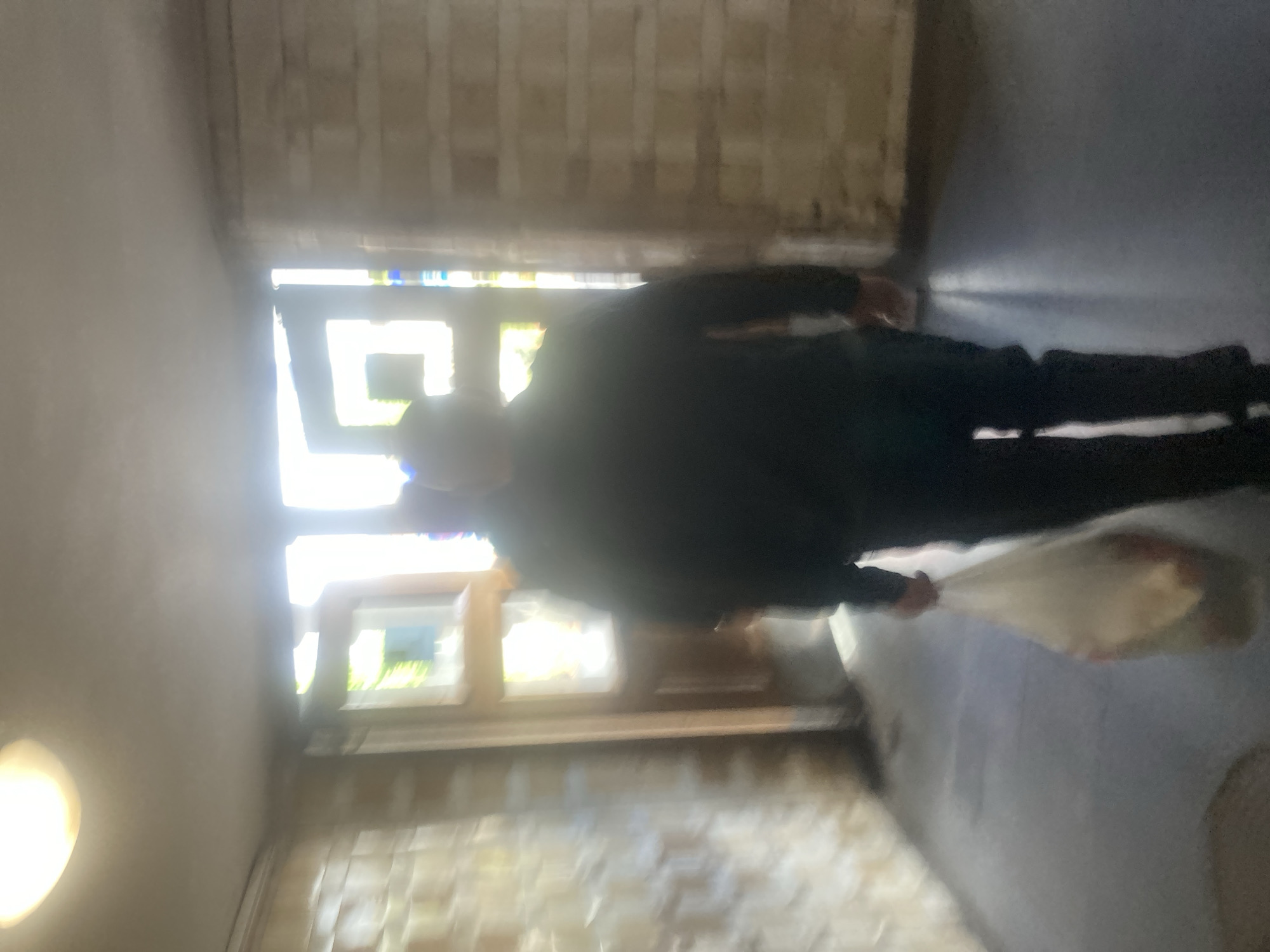 A person in dark clothing stands in a hallway with patterned brick walls and multiple windows letting in bright daylight. The person holds a white plastic bag. The hallway appears to be inside a building, illuminated by a light fixture on the left. The location is possibly in Moscow, Russia.