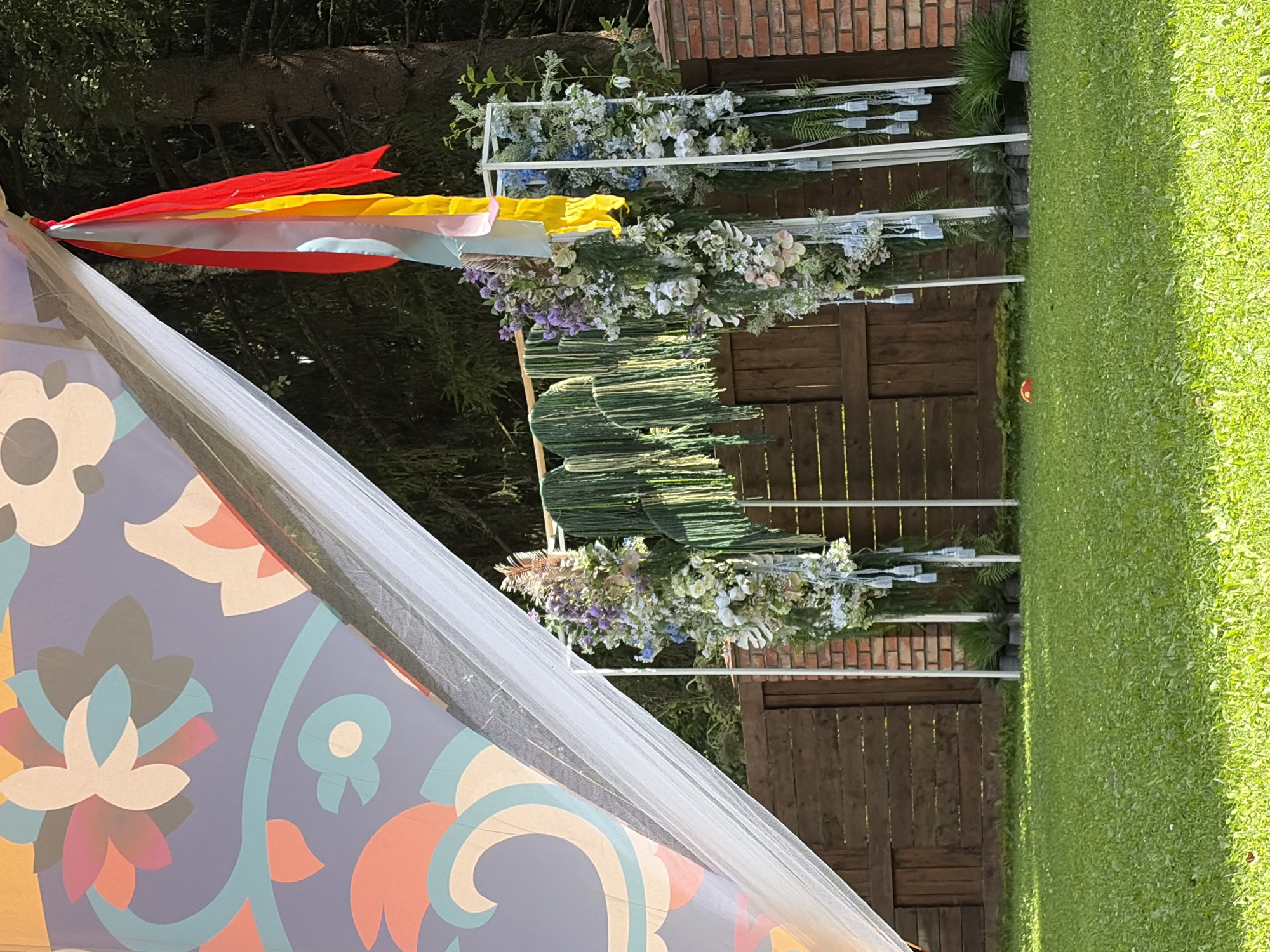 This image captures an outdoor wedding arch in Shebanovo, Russia. The arch is decorated with lush greenery and white and purple flowers, and hangs with tassels and ribbons of various colors. It is situated in a grassy yard, with a wooden fence and brick wall in the background. A large, colorful canopy with a floral pattern is visible in the upper left corner of the image. The scene appears to be set up for an event, possibly a wedding or a festival.