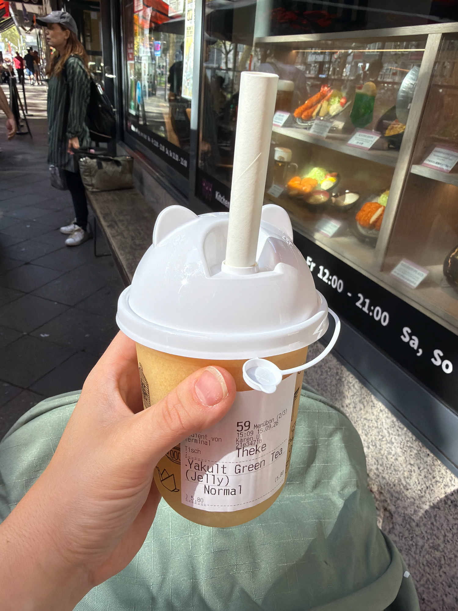 The image shows a person holding a beverage in a white cup with a novelty lid resembling cat ears. The cup has a label with German text that appears to be order information, including a date stamp of "15.09.26". The beverage is a yellow-green color and has a straw made of rolled paper. In the background, a storefront window displays various food replicas, and signage indicates operating hours: "Fr 12:00 - 21:00 Sa, So". A woman wearing a cap and striped shirt is visible in the background standing on a bench. The overall setting suggests an outdoor urban environment, likely in Dusseldorf, Germany, given the common use of German in such establishments.