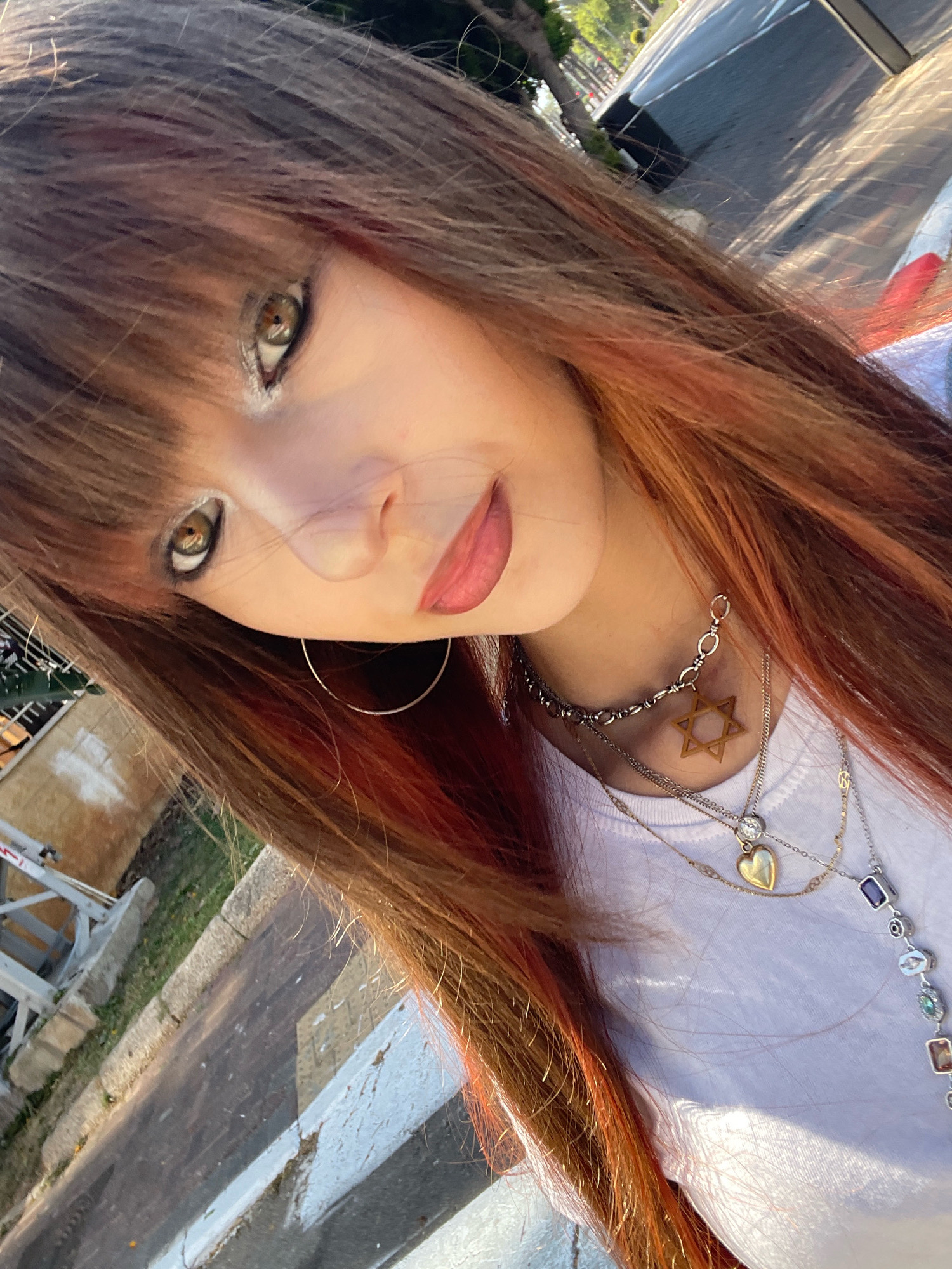 A young woman with long, reddish-brown hair and bangs is pictured in a close-up selfie. She is wearing a white t-shirt and multiple necklaces. One necklace features a Star of David pendant. Her eyes are light brown, and she is wearing makeup. The background appears to be an outdoor urban setting, possibly Raanana, Israel, with a paved surface, some greenery, and a car visible in the upper right corner. The lighting suggests it is daytime.