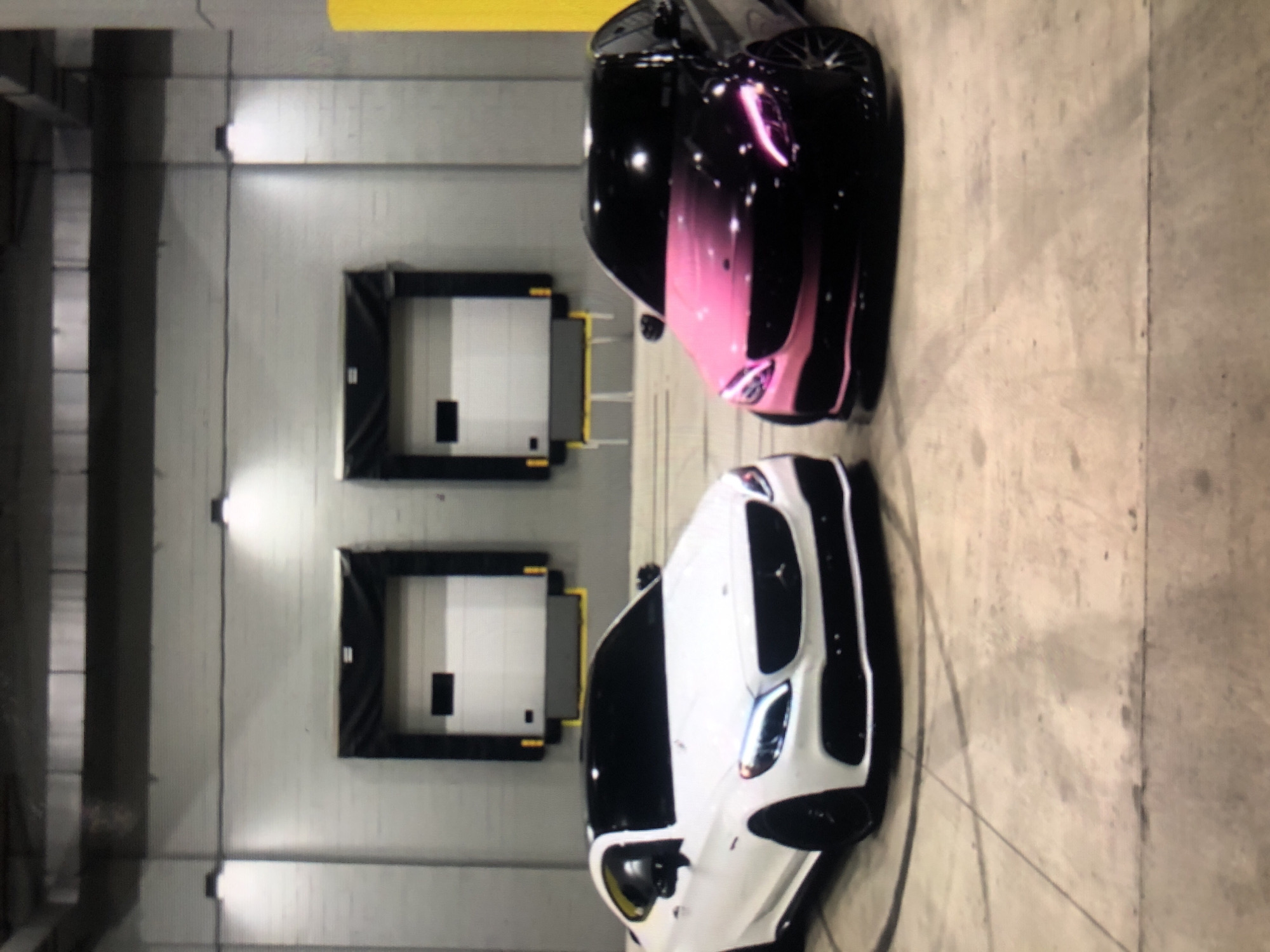 The image shows two cars parked in an indoor parking area. The parking area has grey walls with two loading dock doors. The car closer to the viewer is white, while the other car is black with a pink stripe. The location is unknown and there is no indication this is Lhokseumawe, Indonesia. There are no visible people.