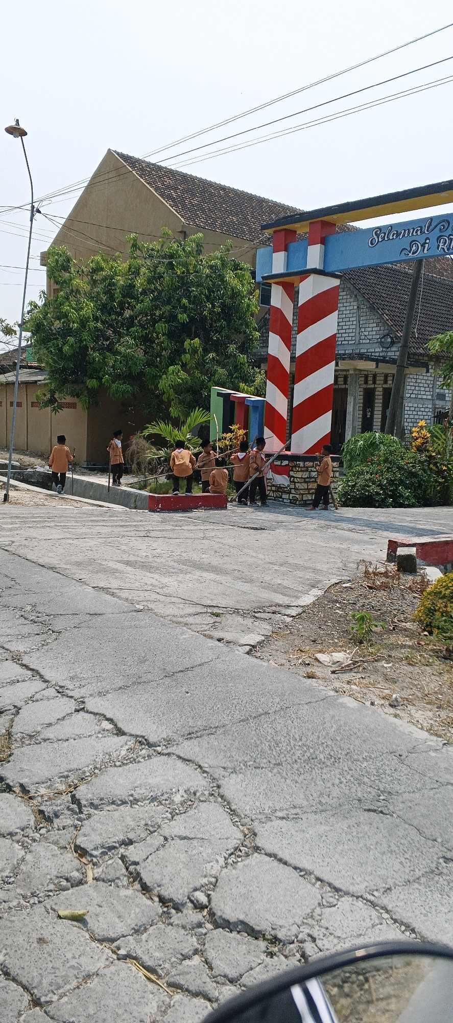 This image shows a group of young people, likely students in uniform, gathered near an archway that reads "Selamat Datang di RI". The archway is decorated with red and white stripes and appears to be an entrance to a locality. The students are holding long sticks and seem to be engaged in some activity together, possibly cleaning or participating in a group exercise. They are on a cracked asphalt road. In the background, there are houses with tiled roofs and lush green trees. A street lamp is visible to the left. The sky is clear and bright. The scene is set in the city of Latukan, Indonesia.
