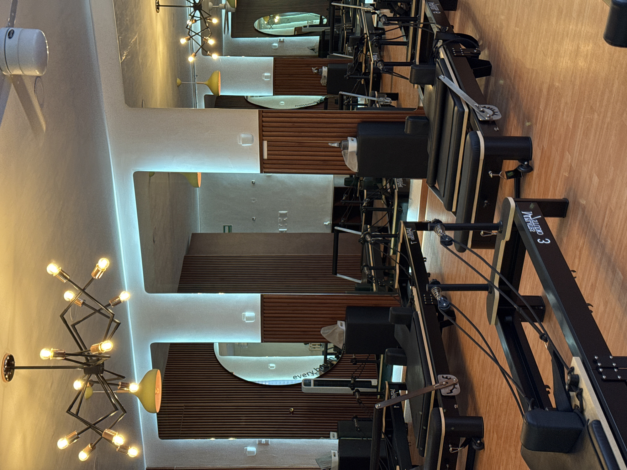The image shows the interior of a pilates studio in Puebla, Mexico. Several Pilates Reformer machines are arranged in a row on a wooden floor. The walls are vertically paneled with dark wood, interrupted by sections of white paint. Mirrors are mounted on the walls. A modern chandelier with multiple exposed bulbs hangs from the ceiling.