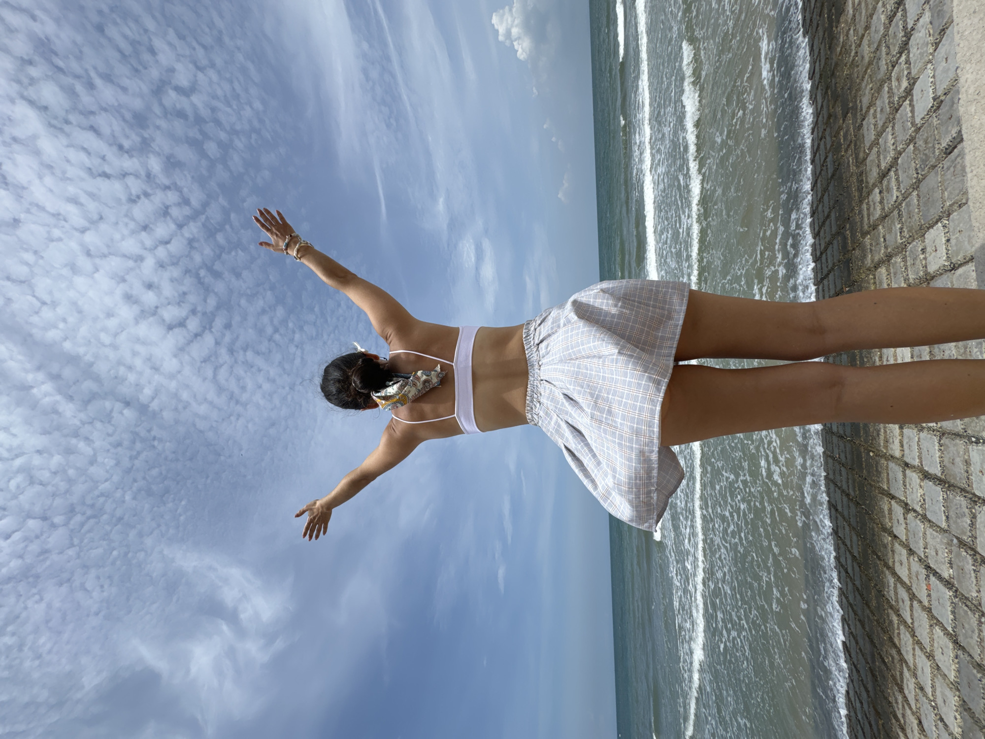 The image depicts a woman with her back to the camera, standing on a paved walkway with her arms outstretched towards a blue sky filled with scattered clouds. She is wearing a white bikini top and a short, light-colored plaid skirt that appears to be caught by the wind. A patterned scarf is tied around her bun. The ocean is visible behind her, with gentle waves rolling towards the shore. The location is identified as Mũi Né, Vietnam.