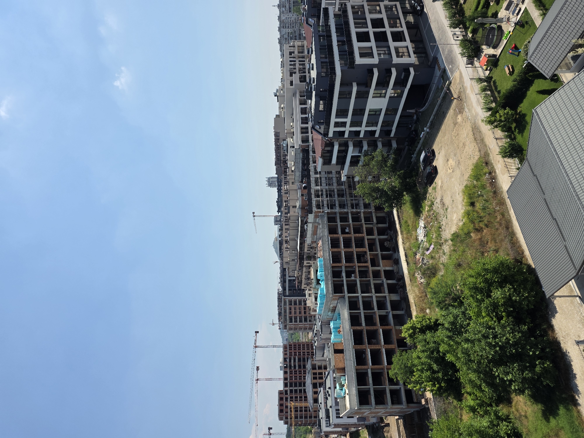 The image depicts an aerial view of a residential construction site in Plovdiv, Bulgaria, on a clear, sunny day. In the foreground, a modern building with a dark facade and white accents is partially visible, with its upper floors featuring balconies. Adjacent to it, a building under construction shows its concrete frame and brickwork, with stacks of blue insulation material on its roof. Several other multi-story buildings, some in various stages of construction with visible cranes, dominate the middle ground. The background reveals a cityscape with more buildings and a distant hill. The site includes areas of bare earth, green vegetation, trees, and a small playground with a trampoline, picnic tables, and colorful play structures in the lower right quadrant. Two cars are parked on a dirt road near the construction site. A lone worker is visible raking the ground near the playground. The overall scene suggests ongoing development in a suburban or semi-urban area of Plovdiv.