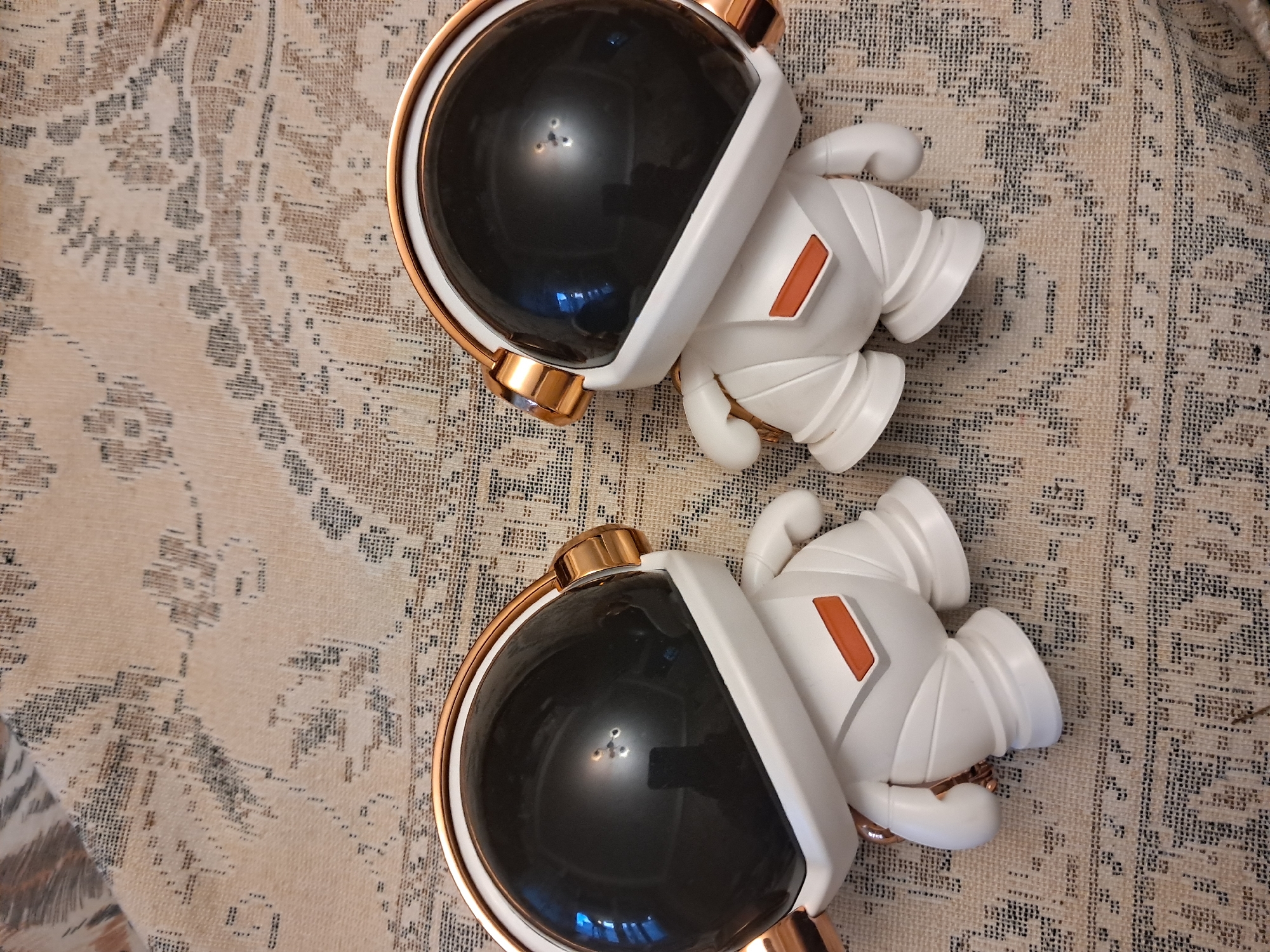 Two white astronaut figurines with black helmets and copper accents are positioned on a patterned rug. The figurines are facing towards the right of the frame, with their helmets tilted downwards. The rug features a repeating geometric pattern in shades of beige and dark gray. The lighting suggests an indoor environment, possibly a home or studio, with reflections visible on the helmets of the figurines.