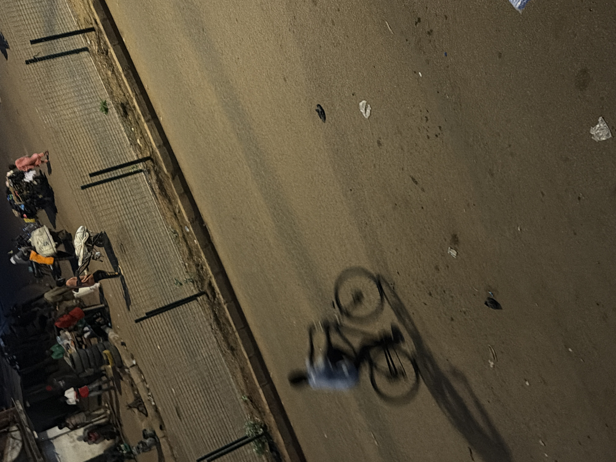 This is an outdoor, ground-level, eye-level, medium shot of a street scene in Bukuru, Nigeria. In the foreground, a person is riding a bicycle, the movement captured with motion blur. The cyclist is wearing a light blue short-sleeved shirt and dark pants. The bicycle has a dark frame and its wheels are blurred due to movement. The street is paved with asphalt and appears somewhat worn, with scattered debris and litter. A long shadow of the cyclist and bicycle is cast on the street. In the middle ground, there is a fence made of wire mesh supported by dark posts, separating the street from a sidewalk or adjacent area. Beyond the fence, several people are visible in the background. Some are standing near what appears to be a market stall with a table laden with goods, including stacked tires and various items. One person is wearing an orange garment, and others are in casual clothing. There are also carts and a motor vehicle present. The lighting suggests it is either dusk or nighttime, with artificial lights from the market area illuminating the scene. The overall atmosphere is that of a busy, possibly commercial, street in an urban environment.