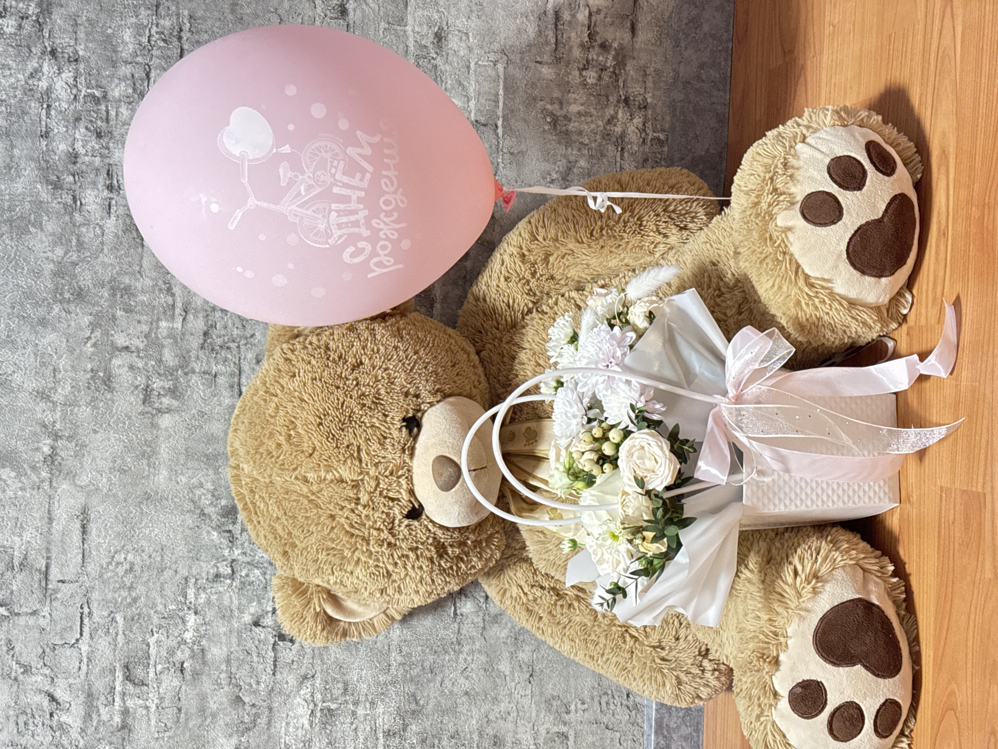 A teddy bear lies on the floor, holding a bouquet of white flowers in its paws and a pink balloon tied to it. The bear is positioned on the border between a light gray, textured surface and a wooden floor. The balloon features a bicycle and Cyrillic text, likely wishing someone a happy birthday. The bouquet is arranged in a white container with white and pale pink ribbons. The bear's paw pads are dark brown. The overall scene appears to be celebratory.