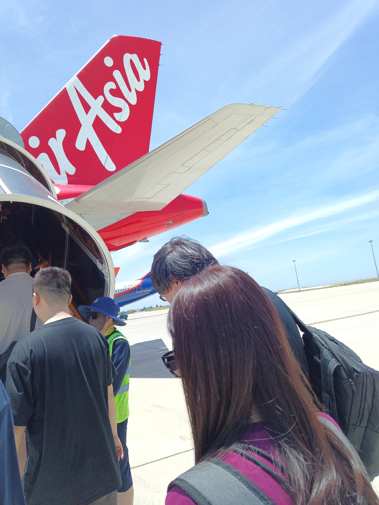 The image shows a group of people boarding an Air Asia airplane on the tarmac at Cam Ranh Airport, Vietnam. The tail of the Air Asia plane, with its distinctive red and white logo, is visible against a clear blue sky. Several passengers are seen from behind, walking towards the open aircraft door. One passenger is wearing a black t-shirt, while another, closer to the foreground, has long brown hair. A person wearing a high-visibility vest and a blue hat, possibly an airport ground staff member, is also present. In the background, another airplane, an Airbus A320 operated by a different airline, is parked on the tarmac. The airport environment is characterized by a paved surface and distant lighting poles under a bright, sunny sky.