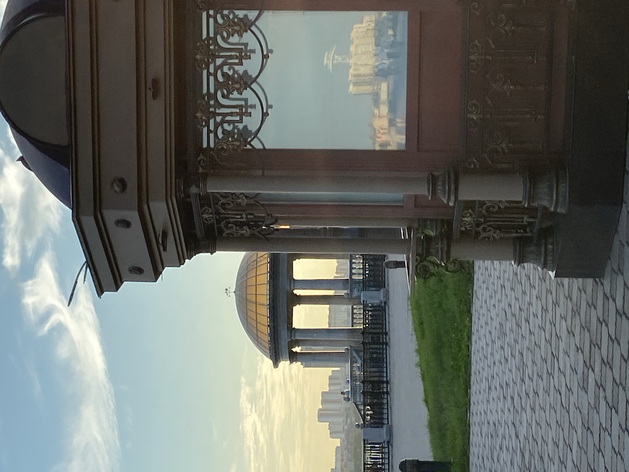 The image depicts an outdoor scene with architectural elements and a cityscape in the background. In the foreground, to the right, is a portion of a dark, ornate structure, possibly a pavilion or kiosk, with intricate metalwork and a large reflective surface. To the left and center of the image, a domed rotunda with a golden roof is visible, supported by columns. It appears to be a public space with a paved walkway in the foreground and a grassy area behind it. A metal railing separates the walkway from the area with the rotunda, beyond which a city skyline with numerous buildings can be seen. The sky is a soft blue with wispy clouds. The reflection in the ornate structure's window shows buildings and people, suggesting this is likely a daytime scene. The scene is identifiable as being located on the embankment in Blagoveshchensk, Russia, looking across the Amur River towards China.