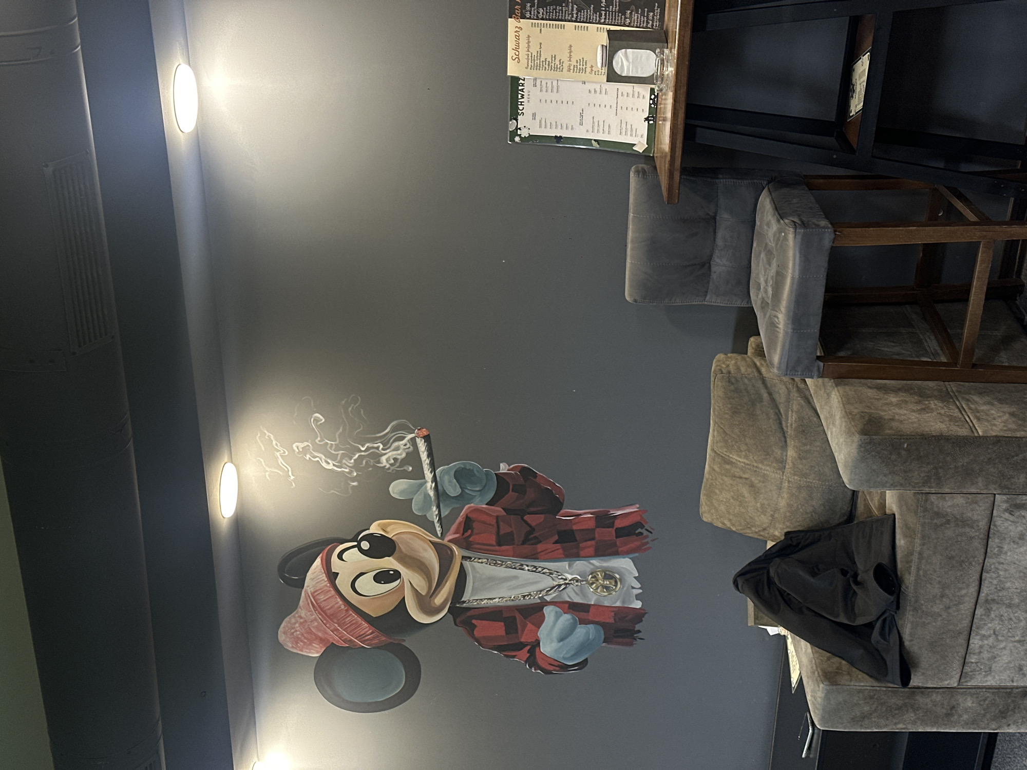 The image depicts the interior of a restaurant or bar in Yerevan, Armenia. A menu titled "Schwarz bar" is visible on a wooden table near gray chairs. A wall mural of Mickey Mouse smoking a cigarette is present, illuminated by nearby wall lights. A gray sofa with a black jacket on it is positioned in the foreground. The environment has a modern, urban aesthetic.