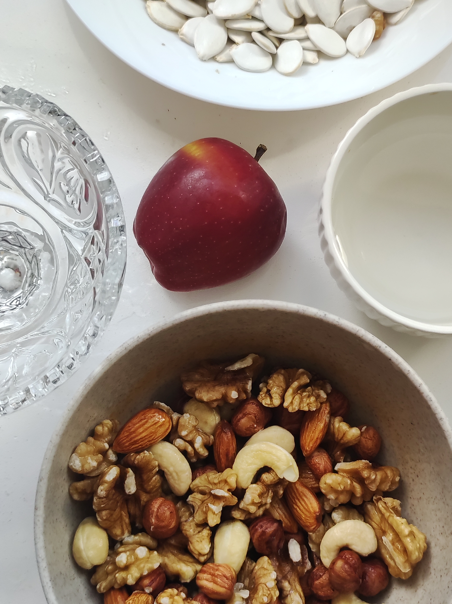 An overhead shot shows a collection of healthy snacks arranged on a white surface. In the foreground, a textured bowl overflows with a mix of nuts including almonds, walnuts, cashews, and hazelnuts. To the upper left, a cut glass bowl is partially visible, appearing to hold water. In the center of the image, a single, vibrant red apple rests on the white surface. To the upper right, a small white ceramic bowl sits empty, its rim decorated with a subtle fluted pattern. Behind the apple and ceramic bowl, a white plate is filled with a pile of light-colored pumpkin seeds. The overall impression is one of simple, wholesome food items presented in an organized, clean setting.