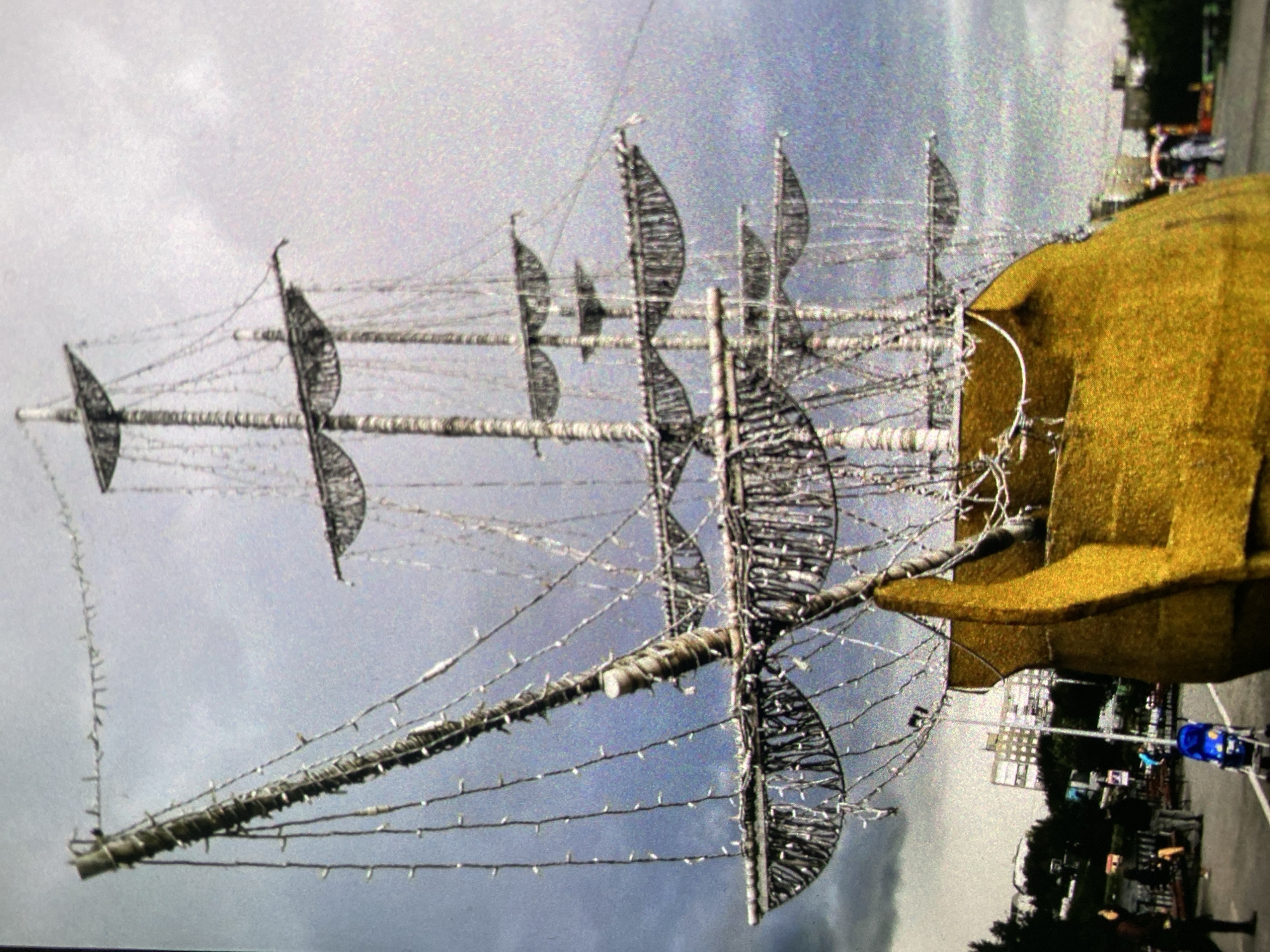 The image shows a decorative ship structure. The ship has masts and sails made of what appears to be wire or light strands, with miniature lights lining the ropes and masts. The hull is covered in a yellow material. In the background, there are trees, buildings, and people, suggesting the ship is located in a public space. Based on the ship's style and the presence of recognizable architecture in the background, it's likely located in Saint Petersburg, Russia. The day is overcast, indicated by the gray sky. There are people walking nearby.