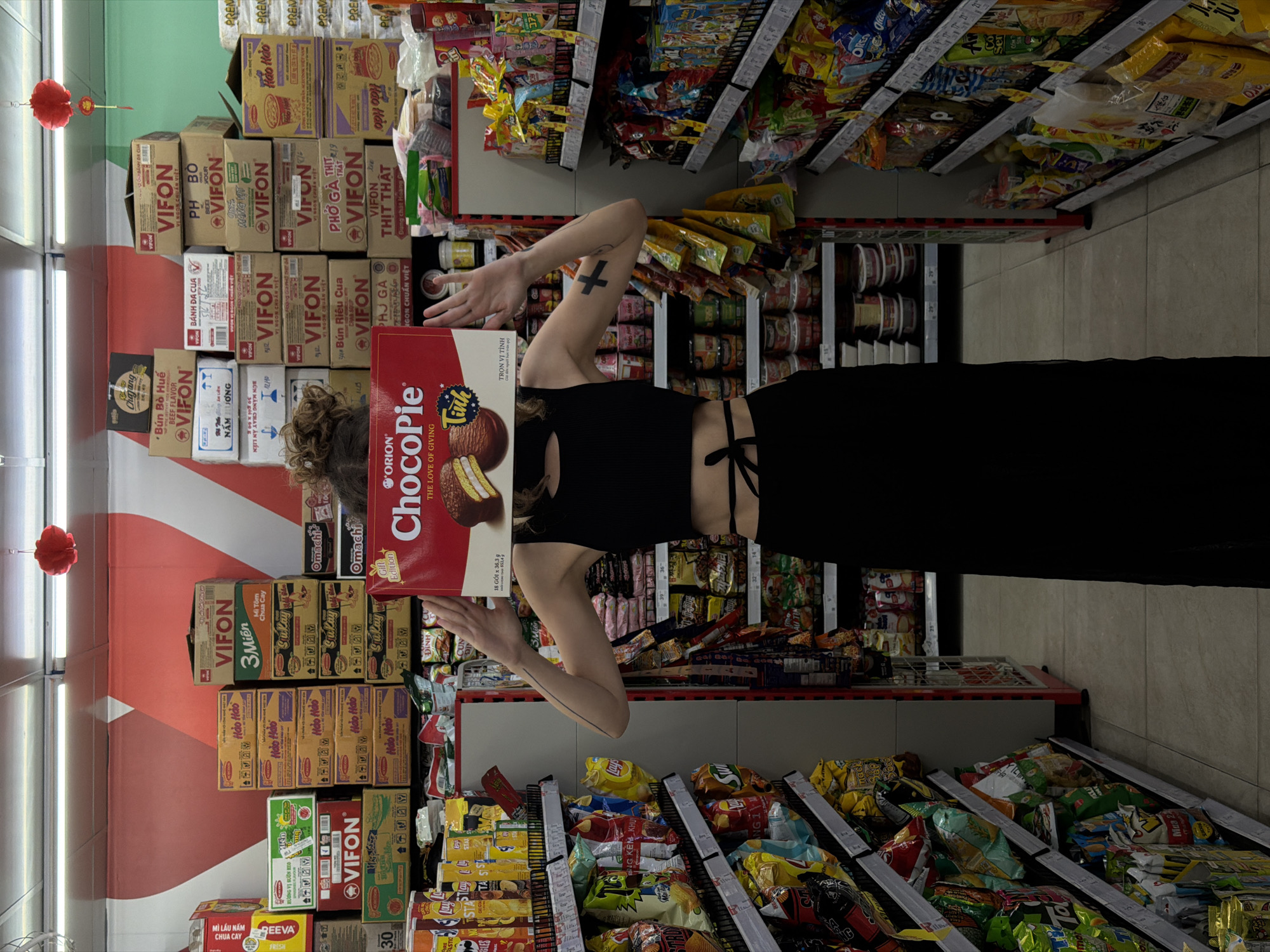 A person stands in a supermarket aisle, holding a large red box of "ORION ChocoPie" directly in front of their face, completely obscuring it. The person is wearing a black sleeveless crop top and black wide-leg pants. A black cross tattoo is visible on their left arm, and a thin line tattoo on their right forearm.
The background features tall shelves densely packed with various packaged food items, primarily instant noodles and snacks. To the left, a wall displays a red and white diagonal striped pattern, with red ornamental lanterns hanging from the ceiling. Fluorescent lights illuminate the tiled floor and the store interior.
Numerous Vietnamese product brands and texts are visible on the stacked boxes. "VIFON" is prominent with varieties such as "PHỞ BÒ" (Beef Pho), "BÚN RIÊU CUA" (Crab Noodle Soup), "PHỞ GÀ" (Chicken Pho), "THỊT THẬT" (Real Meat), "BÚN BÒ HUẾ" (Hue Beef Noodle), and "MÌ TÔM CHUA CAY" (Spicy Shrimp Noodle). Other notable brands include "Hảo Hảo," often displaying "HƯƠNG VỊ SƯỜN HEO" (Pork Rib Flavor), "Omachi," and "GEEVA MÌ LẨU NẤM CHUA CAY."
The "ChocoPie" box details include "THE LOVE OF GIVING," "Tinh," and "Gift Edition," along with "18 GÓI x 36,3 g" (18 packs x 36.3g). Other visible text fragments on packaging include "Chajang," "AN LIÊN," "Sukay," and phrases like "TRỌN VỊ TÌNH" and "Mua 2 TẶNG 1" (Buy 2 Get 1 Free). Shelf labels display numbers such as "29," "30," "36," "37," and "39." The setting is an indoor grocery store or minimart in Đà Nẵng, Vietnam.
