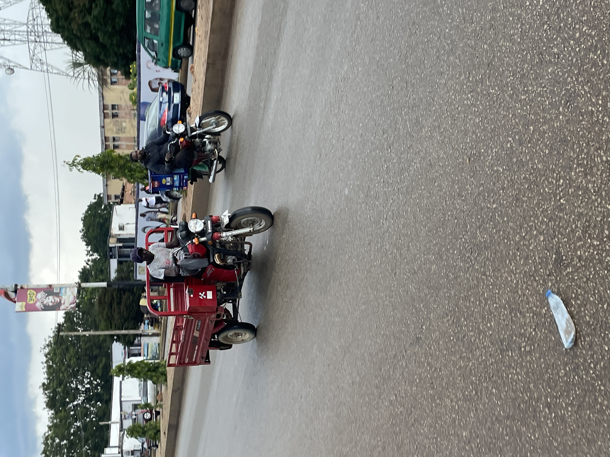 In Jos, Nigeria, two motorized tricycles are visible on a wet asphalt road. The first tricycle, a red cargo model, is in motion, with a person wearing a striped t-shirt and a dark cap operating it. The second tricycle, a motorcycle with a delivery box on the back, is also moving, and its operator is wearing a dark jacket and a dark cap. In the background, buildings, trees, and a cell tower are present. A plastic water bottle lies on the road in the foreground. The sky is overcast.