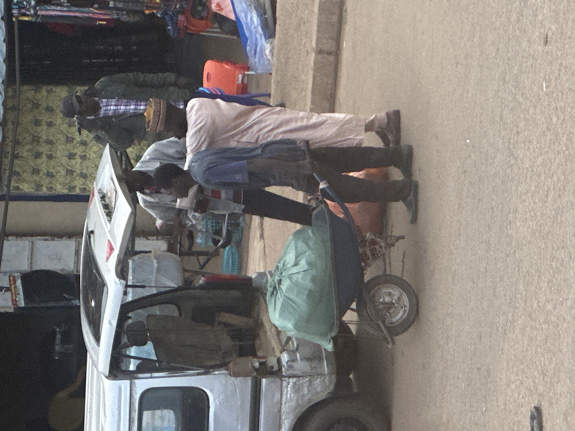 In Bukuru, Nigeria, a scene unfolds in what appears to be a market or roadside area. A silver van, partially open at the rear, is parked on a dirt and asphalt surface. In front of the van, a dark-colored wheelbarrow is positioned, loaded with a large, green, gathered plastic bag. Three men are present. One man, wearing a light-colored long-sleeved shirt and dark pants, is partially visible, facing away from the camera. Another man, in a blue t-shirt and dark pants, is pushing the wheelbarrow, gripping its handles. He appears to be looking forward. A third man, standing to the right of the wheelbarrow, is wearing a light pink traditional garment and a woven hat. He is facing towards the other two men, his left arm raised and holding what appears to be a mobile phone to his ear. In the background, a stall with various items for sale is visible, along with a patterned fabric wall and a dark doorway. The overall lighting suggests daytime.