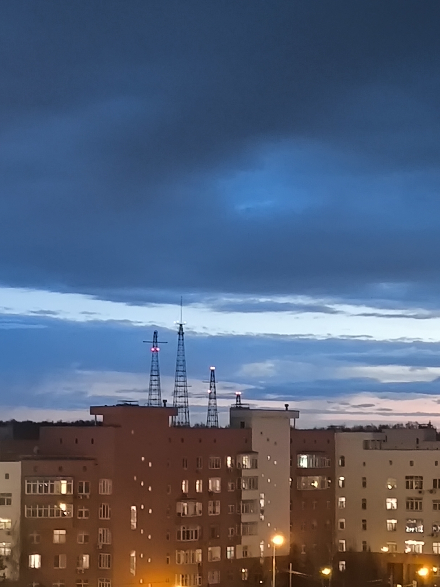 The image shows an urban landscape at dusk in Khimki, Russia. In the foreground are several mid-rise apartment buildings with lit windows. Behind the buildings are three lattice towers, possibly radio or communications towers, marked with red lights. The sky is predominantly blue, with heavy clouds above and a few lighter, thinner clouds near the horizon. The overall lighting suggests it is early evening. There are no visible people or actions depicted in the scene.