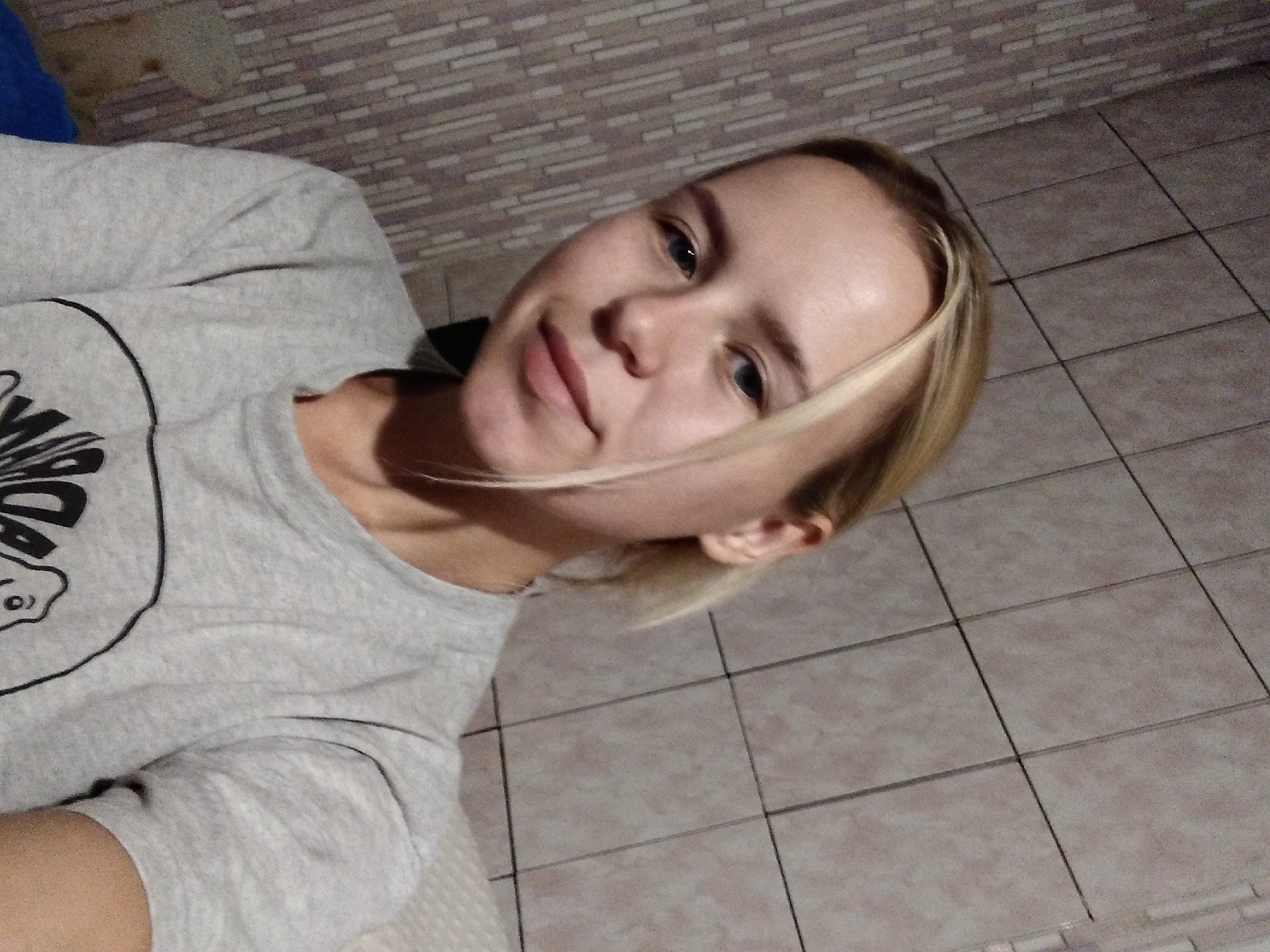 A young woman with blonde hair pulled back into a ponytail is taking a selfie in an indoor setting. She is wearing a grey t-shirt. The background appears to be a bathroom with tiled walls. The lighting suggests it is evening or indoors. No specific details about the city of Radisne, Ukraine, can be determined from the image.