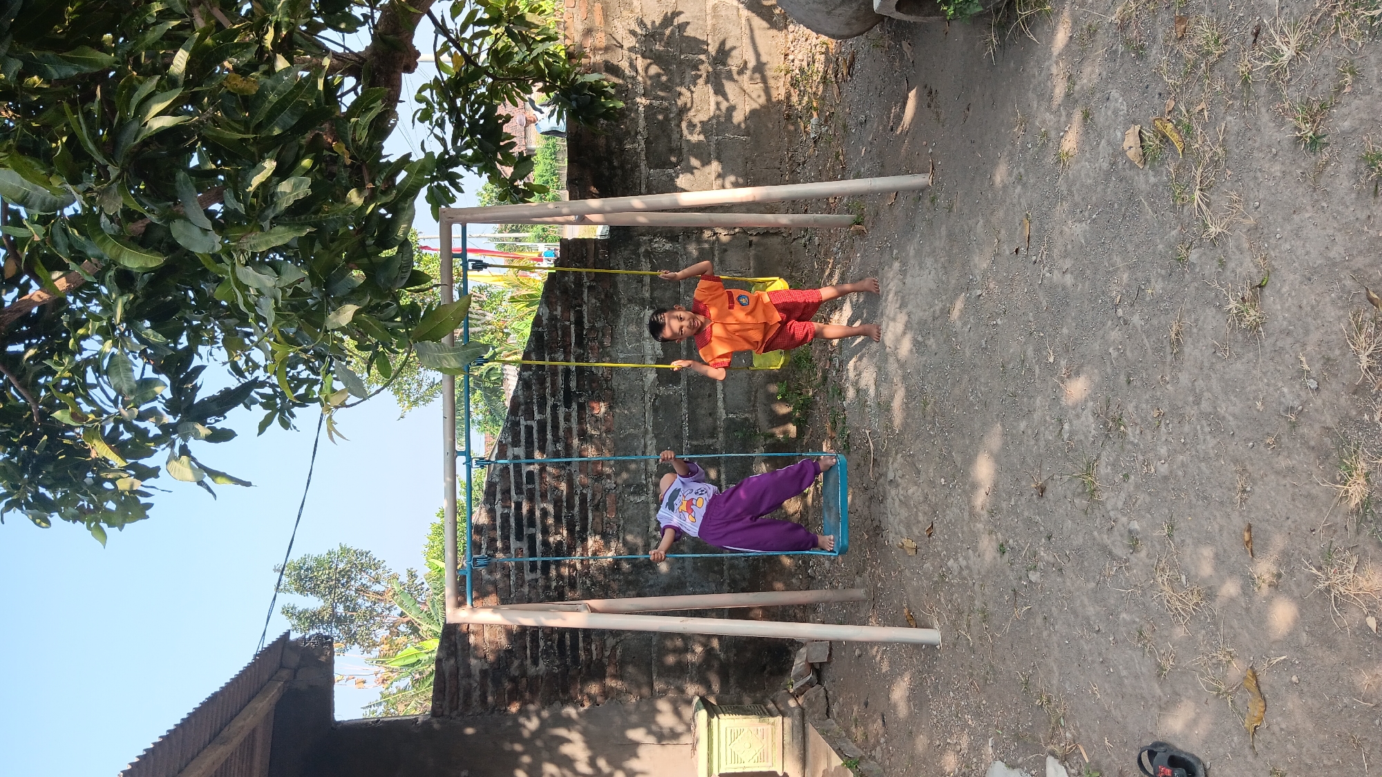 In Sonobekel, Indonesia, two young boys are playing on a swing set. The boy on the left is on a blue swing, wearing purple pants and a light-colored t-shirt with a cartoon character. He is leaning back with his arms outstretched. The boy on the right is on a yellow swing, wearing red shorts and an orange t-shirt. He is holding onto the swing ropes with both hands and looking towards the camera. The swing set is set in a dirt-covered yard, with a brick wall behind it. Trees with green leaves are visible in the background, and the sky is clear and blue. A corrugated metal roof of a building is visible on the left side of the image.