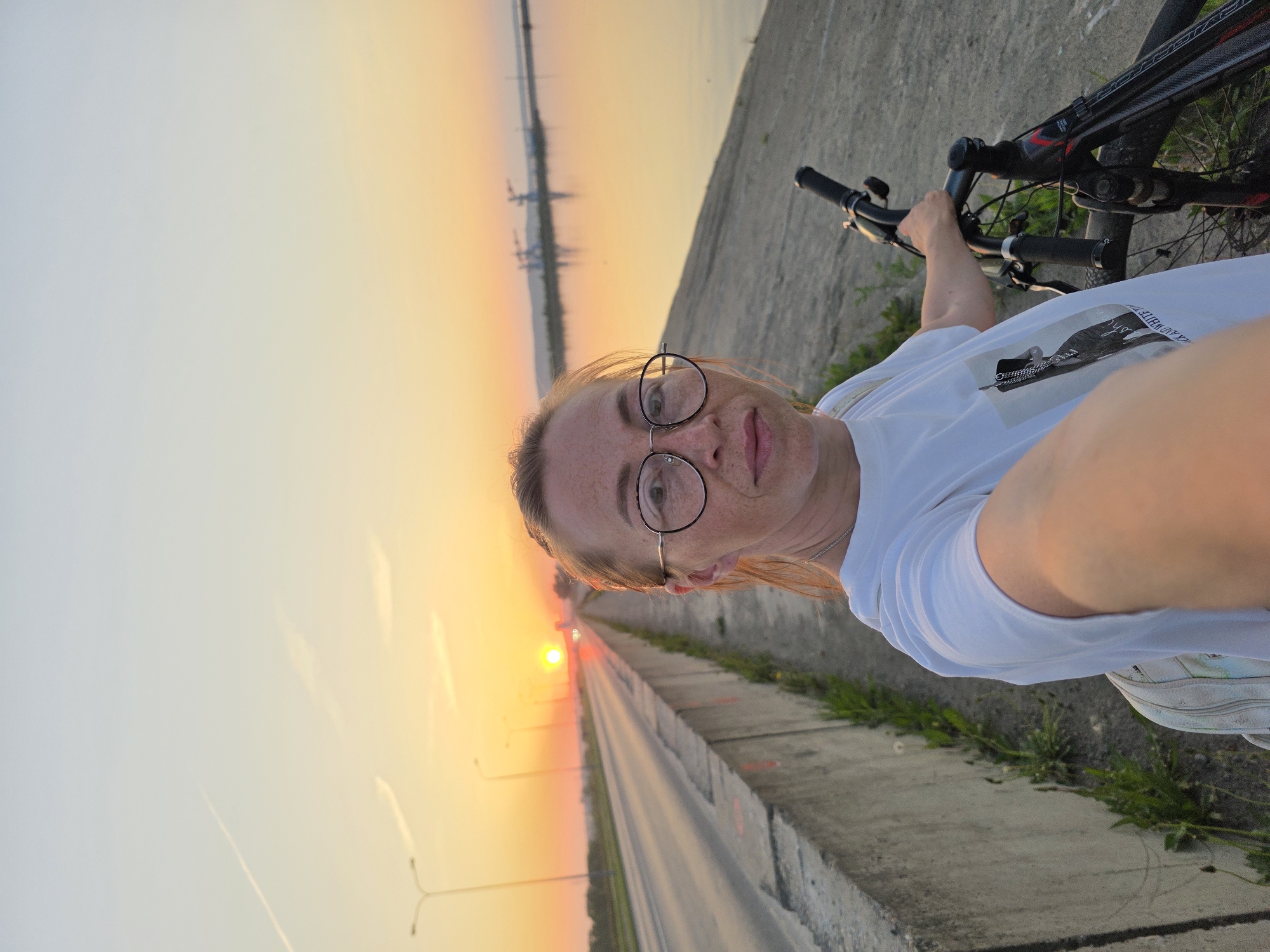 A woman with freckles and glasses is taking a selfie while standing with her bicycle on a concrete path. The sunset is visible in the background, with a road and streetlights leading towards it. The location is the city of Chaikovsky, Russia.