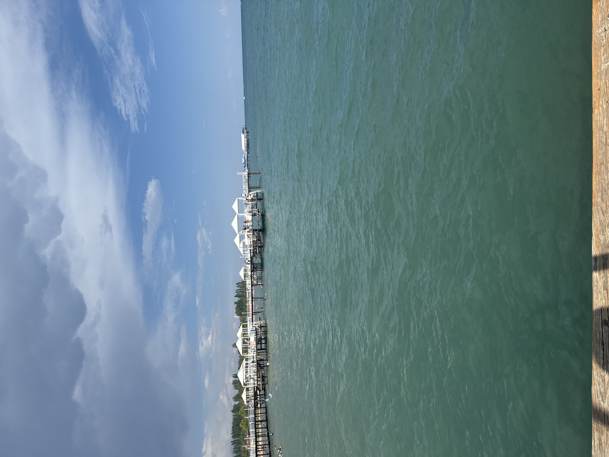 An eye-level outdoor shot captures a body of turquoise water, possibly a lake or sea, in Sary-Oy, Kyrgyzstan. In the distance, a white pier extends into the water, lined with small white canopy-covered structures. People are visible on the pier and in the water, some swimming. The water has a rippled texture. The sky above is a mix of blue and dramatic grey clouds. The foreground shows a wooden surface, possibly the edge of a deck or pier.