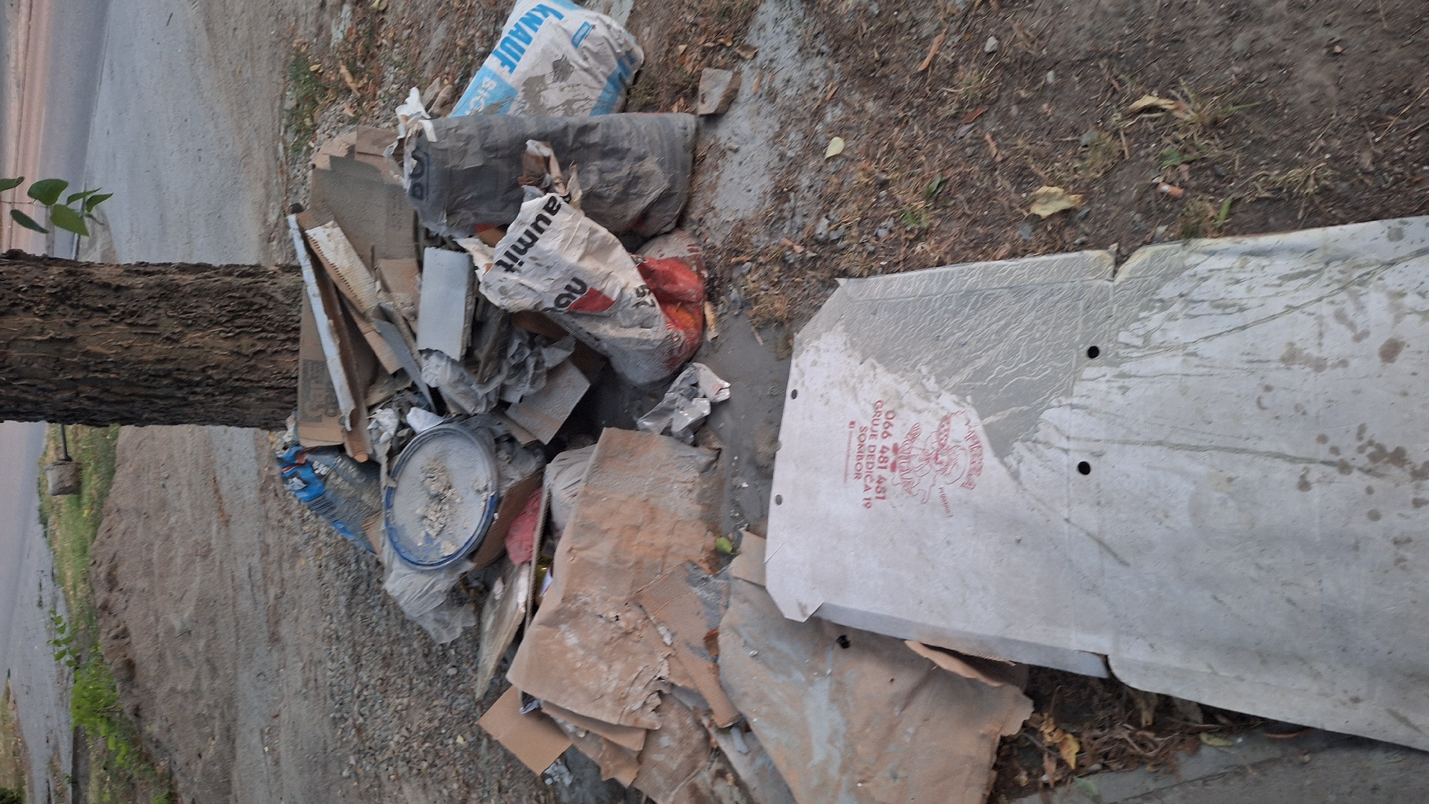 The image depicts a pile of construction debris near a tree on a roadside in Sombor, Serbia. The debris includes cardboard boxes, bags of what appears to be cement or construction mix, plastic sheeting, and broken pieces of tile or similar material. One of the bags is labeled "KNAUF". In the foreground, a large, flat piece of material, possibly a cover or panel, lies on the ground, also showing signs of cementitious material and what appears to be a logo and text in red. The ground is a mix of dirt, gravel, and dry vegetation. The overall scene suggests a temporary storage or dumping area for construction waste in an urban environment.