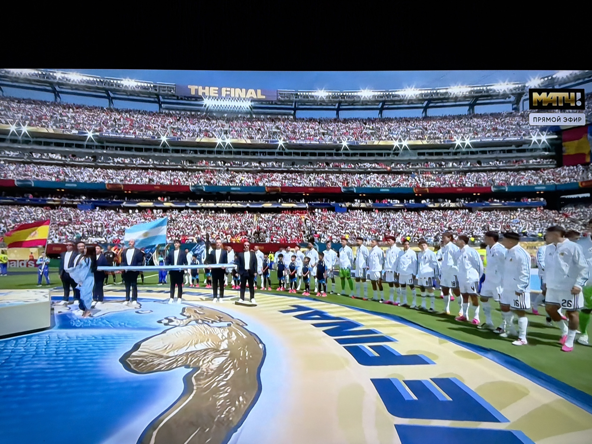 The image shows a wide shot of a football stadium filled with spectators. In the foreground, two teams of players, one in white uniforms and the other in dark blue and black uniforms, stand on the field. A large graphic is present on the field, with the word "FINAL" prominently displayed. Flags of Argentina and Spain are visible in the background. A broadcast logo, "МАТЧ!", with the text "ПРЯМОЙ ЭФИР" (which translates to "LIVE BROADCAST" in Russian) is visible in the upper right corner. There is no specific indication within the image to suggest the location is in Orenburg, Russia.