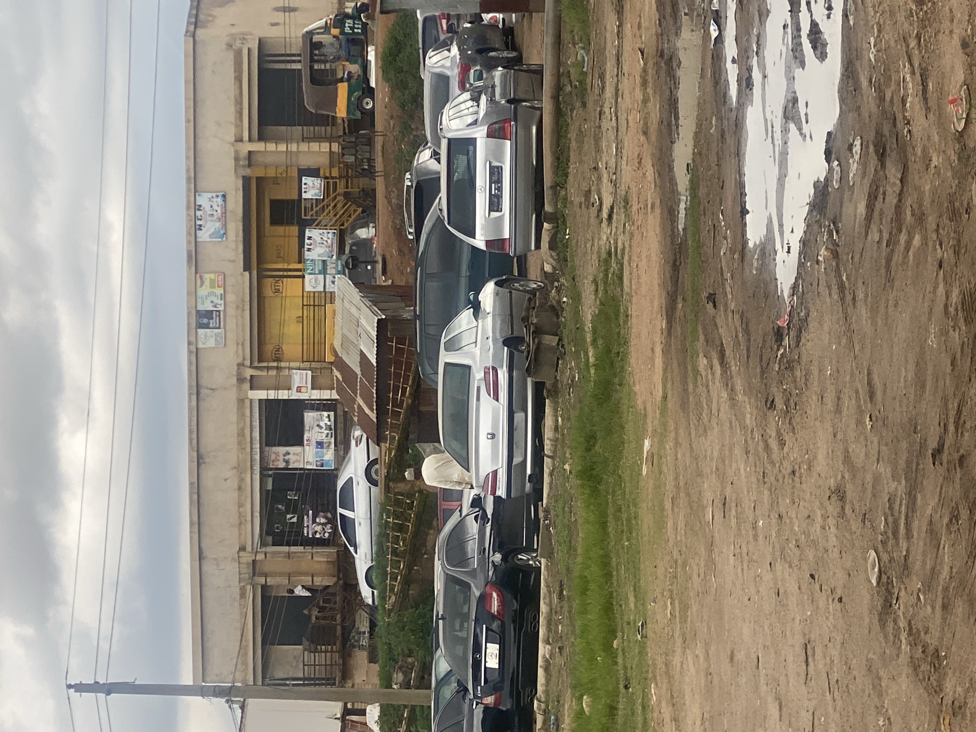 The image depicts an outdoor scene in Rafin Par, Nigeria, likely a commercial area with a building in the background and several vehicles in the foreground. The building appears to be a single-story structure with a light-colored facade and several open storefronts. Signs on the building include "MTN" and "NEN," suggesting telecommunications or related services. A rickshaw-style vehicle is parked to the right of the building, and several cars are parked in the dirt lot in front of the building and to the left. The cars include a black Mercedes-Benz sedan, a silver Peugeot sedan, and a silver Mercedes-Benz station wagon, among others. A man wearing a light-colored robe and a cap is standing between two of the cars. The ground is unpaved, with patches of grass and puddles of water, indicating a potentially unkempt or rural environment. The sky is overcast, suggesting a cloudy day.
