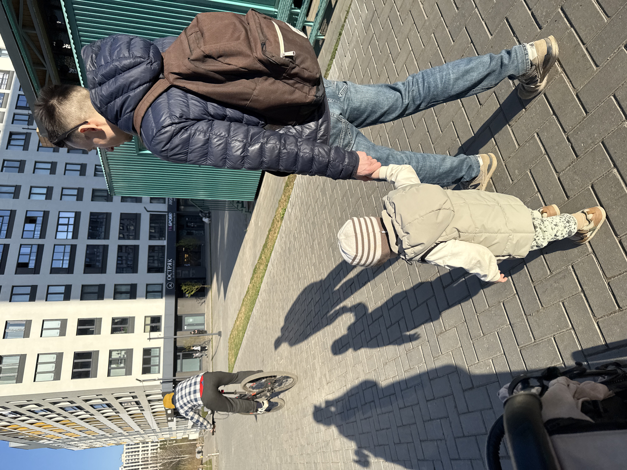 A man with a backpack holds the hand of a child in a striped hat and beige vest. They are walking on a paved sidewalk in front of a modern apartment building with the sign "ОСТРЯК (OSTRYAK)" visible. A cyclist passes nearby. The location is Izhevsk, Russia.