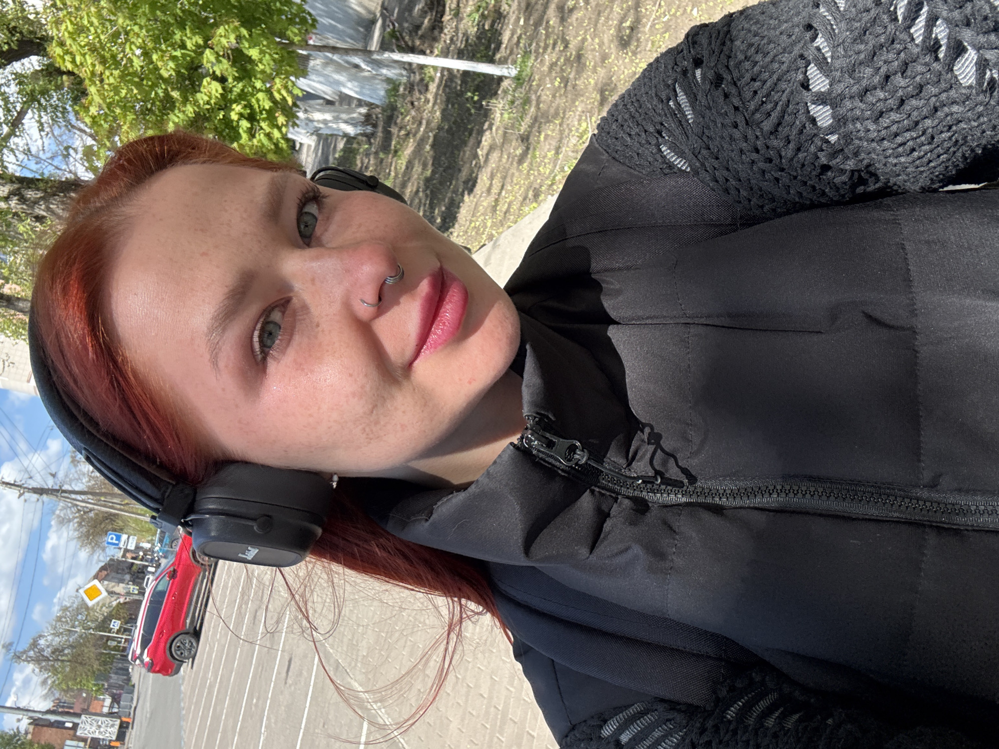 A young woman with red hair, wearing black headphones and a black jacket, takes a selfie outside on a sunny day. She has a nose ring and freckles. In the background, there's a street with a red car, a street sign, and buildings. The location appears to be Rostov-on-Don, Russia, based on the visible street signs.