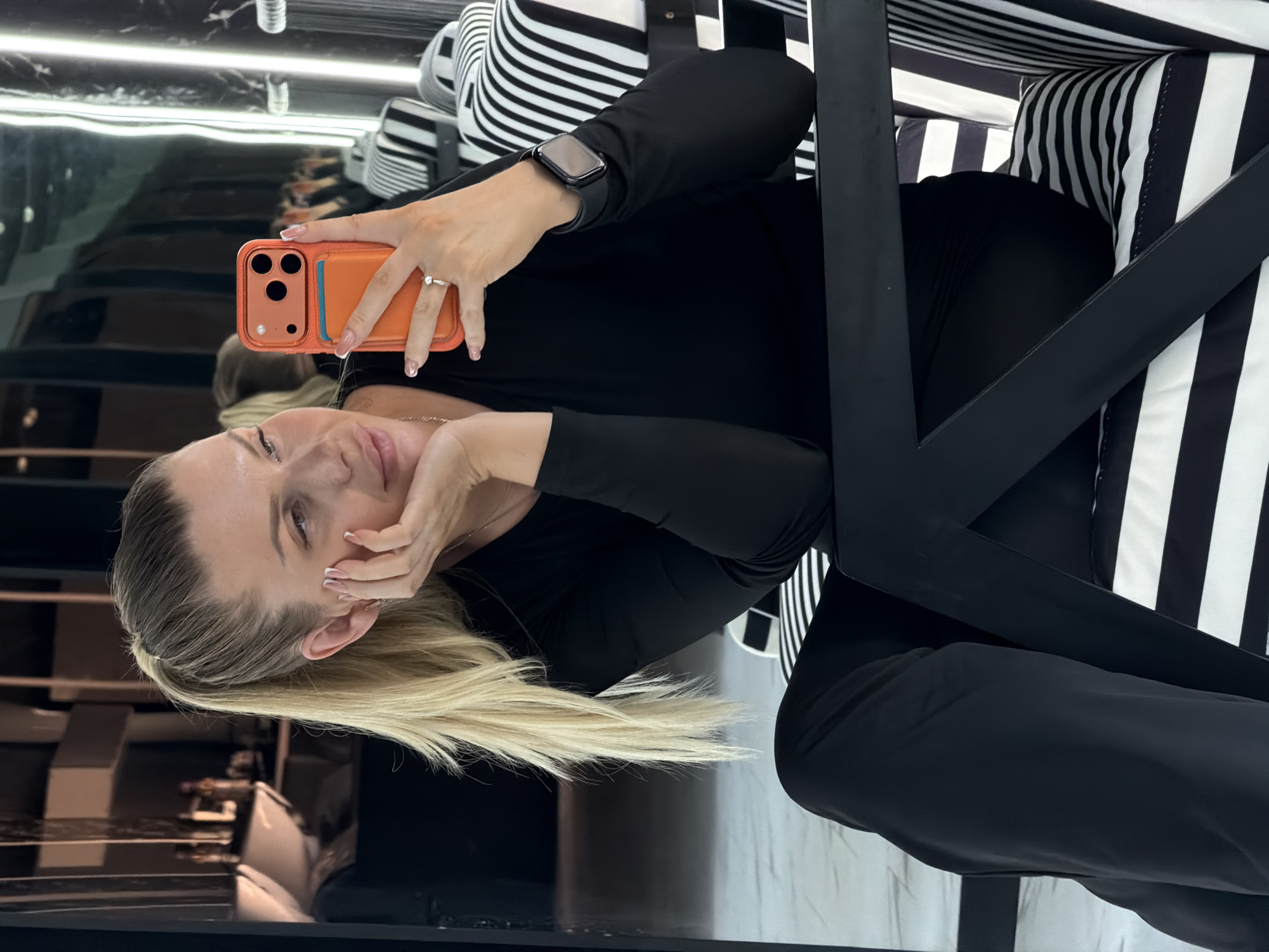 The image shows a woman with blonde hair in a ponytail, wearing a black long-sleeved top and black pants. She is sitting in a chair with black and white striped cushions and holding an orange phone in her right hand, taking a selfie. The background appears to be a room with mirrored walls reflecting shelves with bottles and a person in the background. A portion of the reflection shows a chair similar to the one she is sitting in. The scene suggests an indoor setting, possibly a salon or a modern commercial establishment. There is no clear indication that this location is in Moscow, Russia.